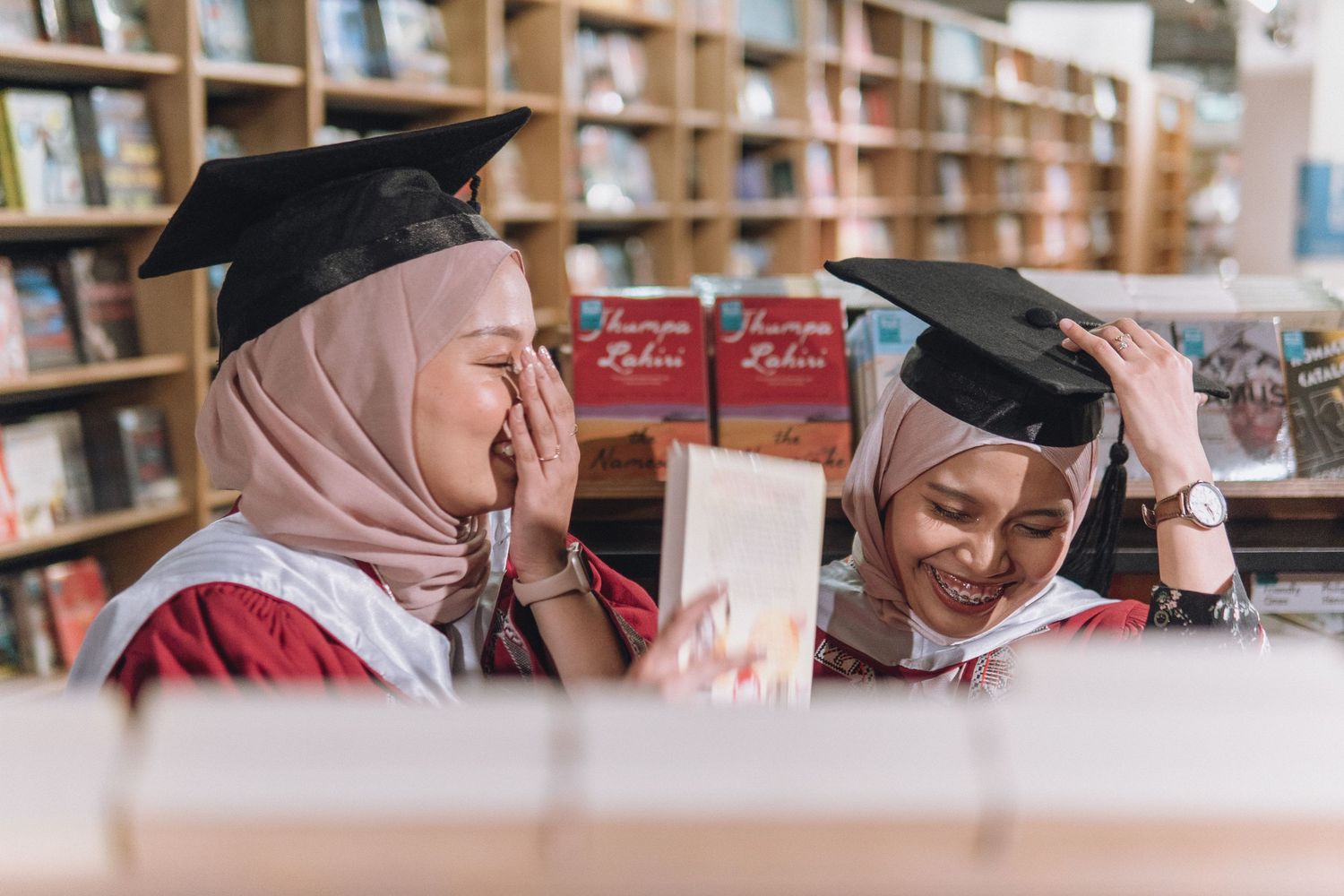 Two graduates in red academic regalia share a joyful moment while looking at books in a library.