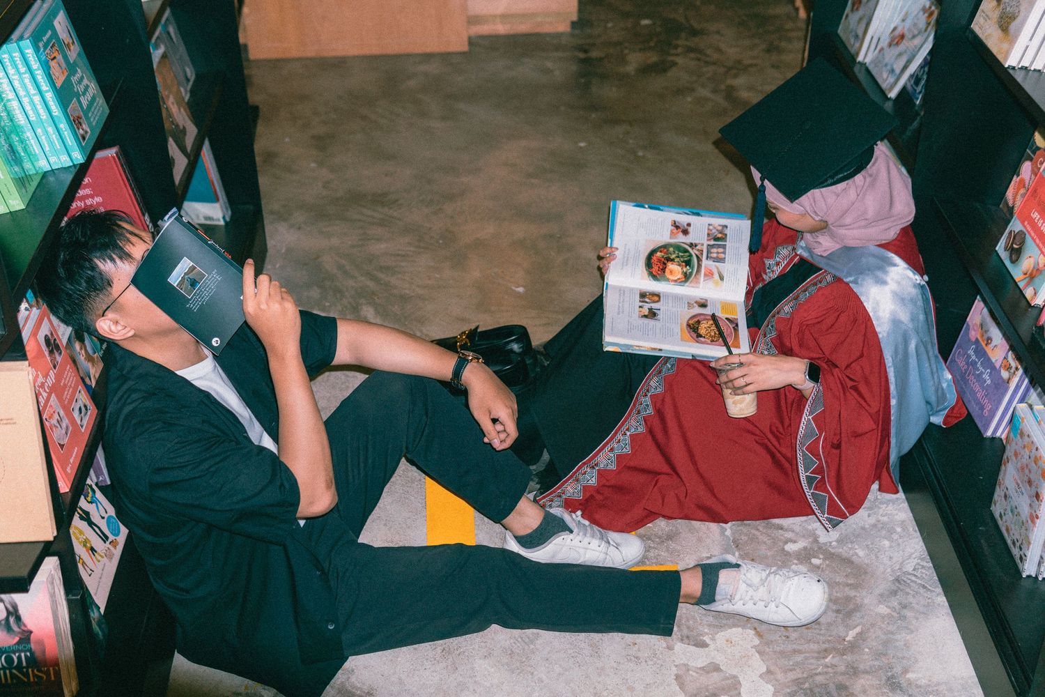 Students in graduation attire sit on the floor of a library while reading books.