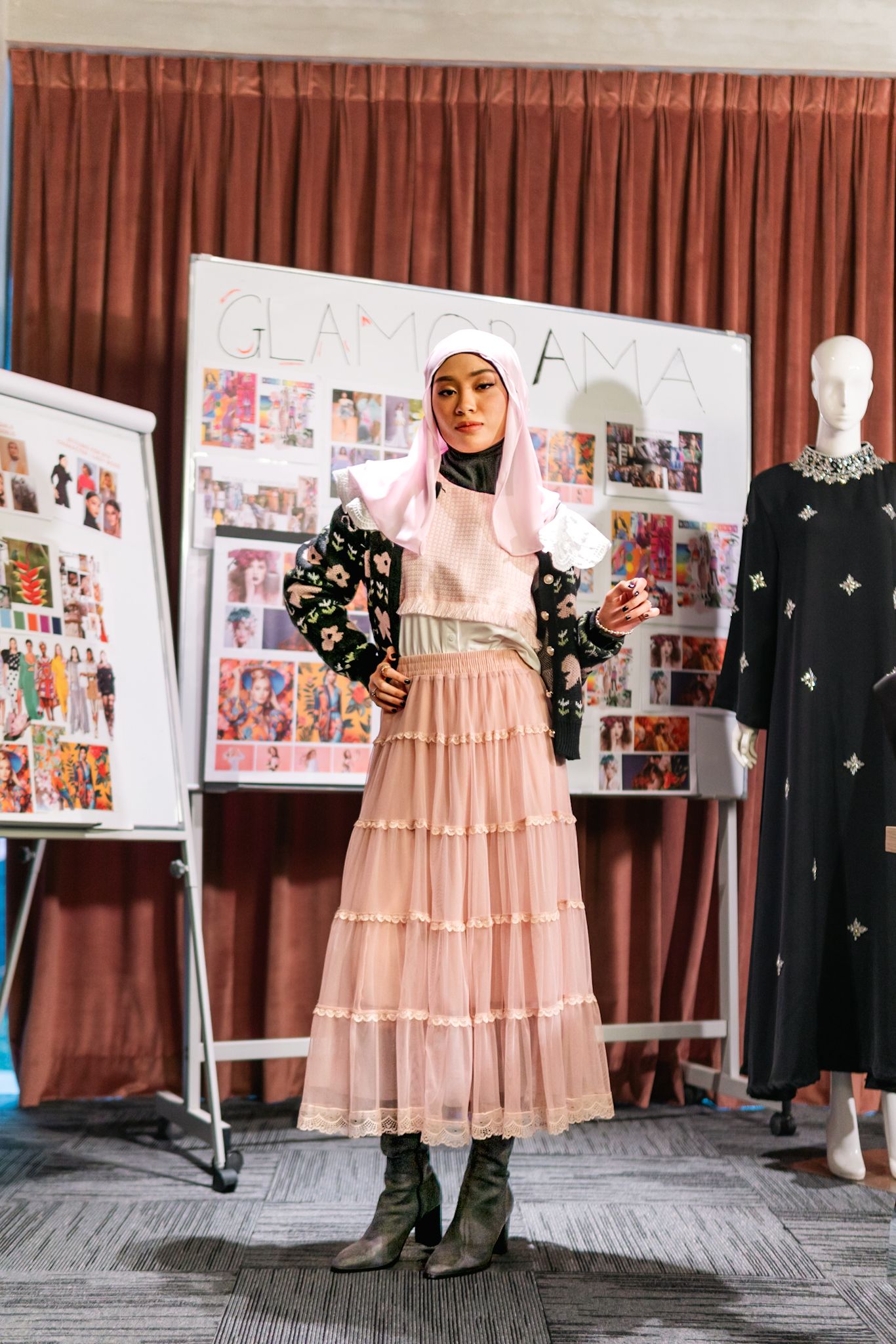 Fashion model wearing pink tiered maxi dress with black floral print jacket and boots in a showroom setting.