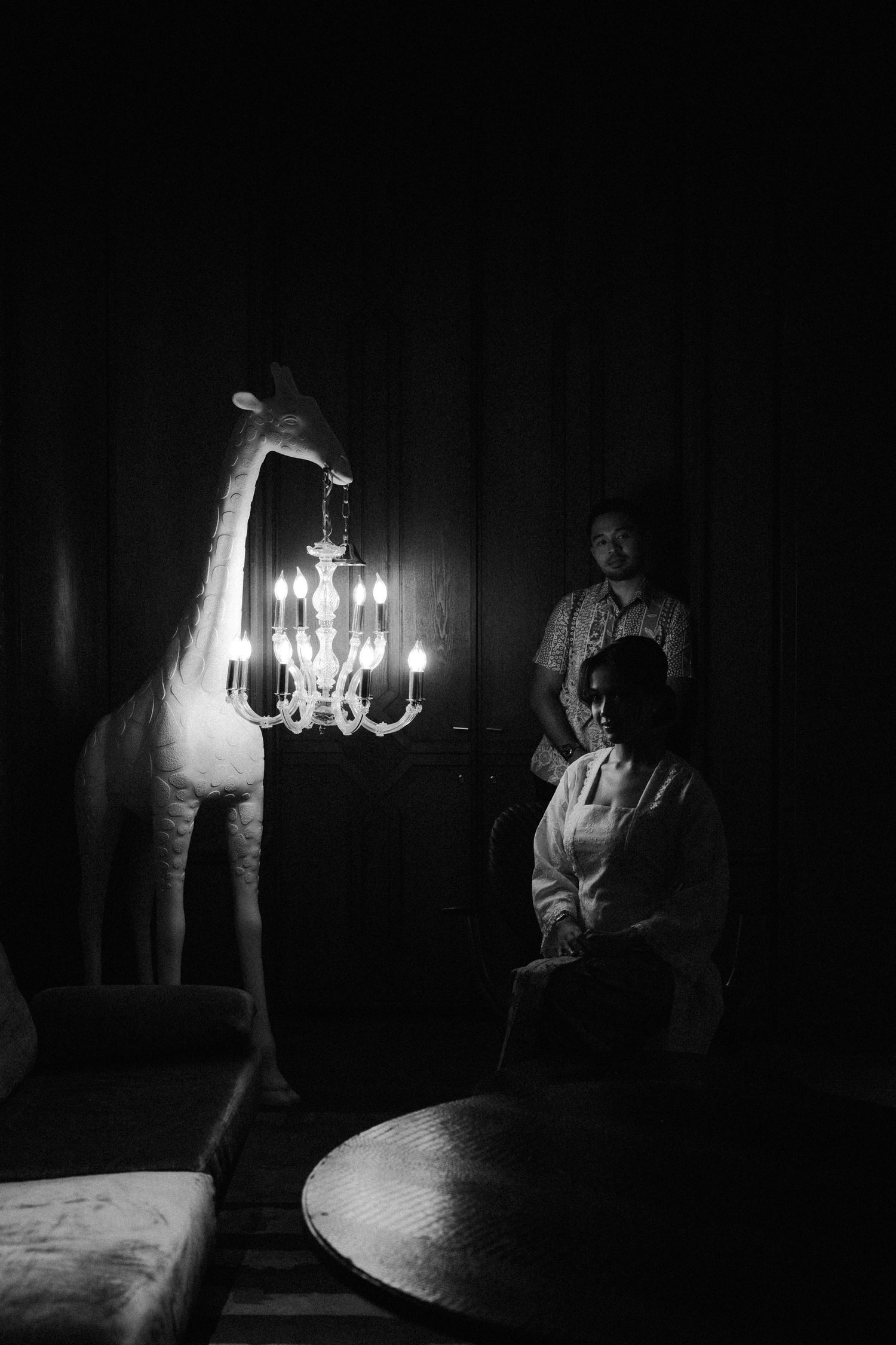 A surreal black and white photo showing a tall giraffe figure next to a crystal chandelier in a darkened room.