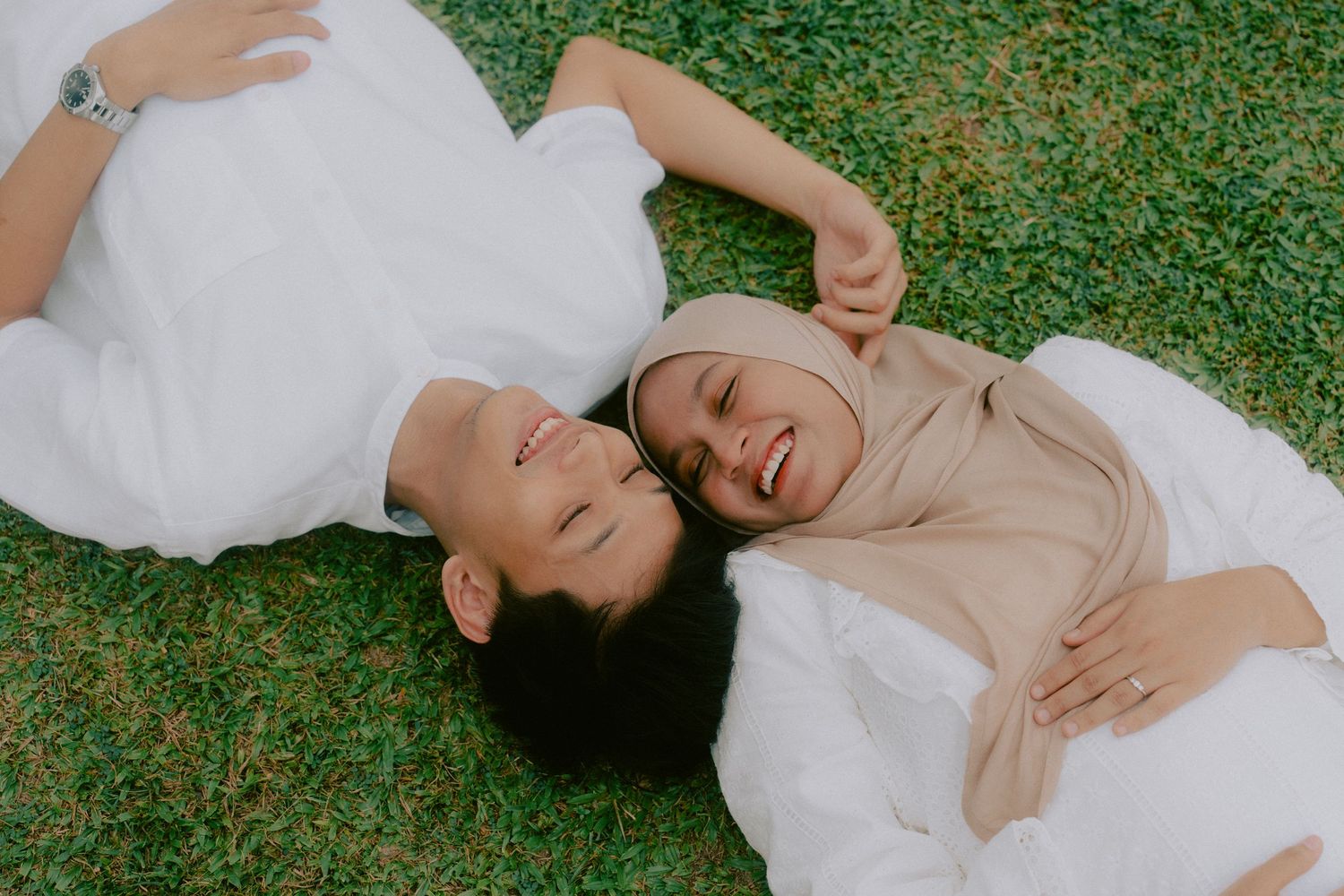 A couple lying on grass wearing white and beige outfits while sharing a happy moment together.