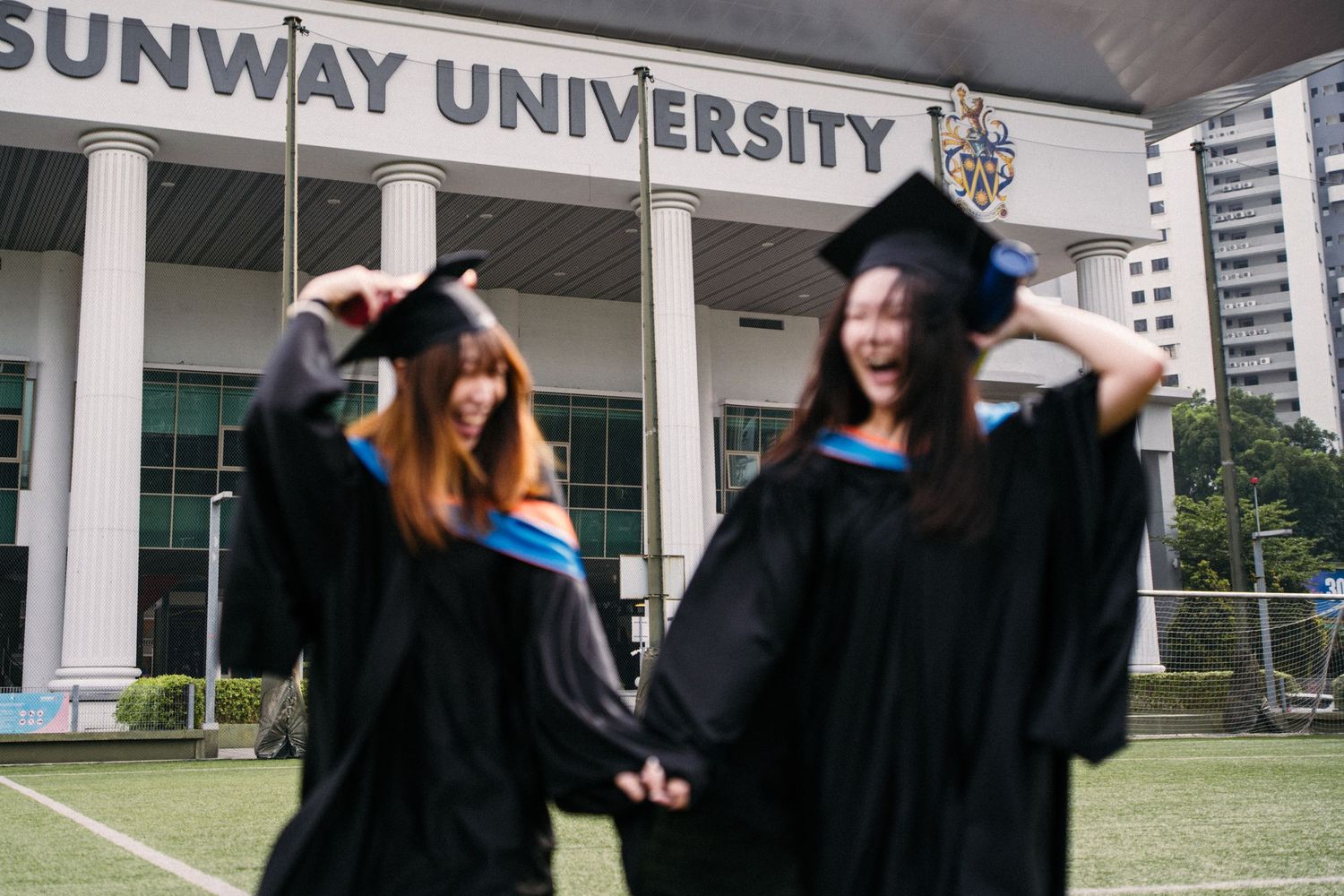 Playful graduates in academic dress posing in front of Sunway University sign.