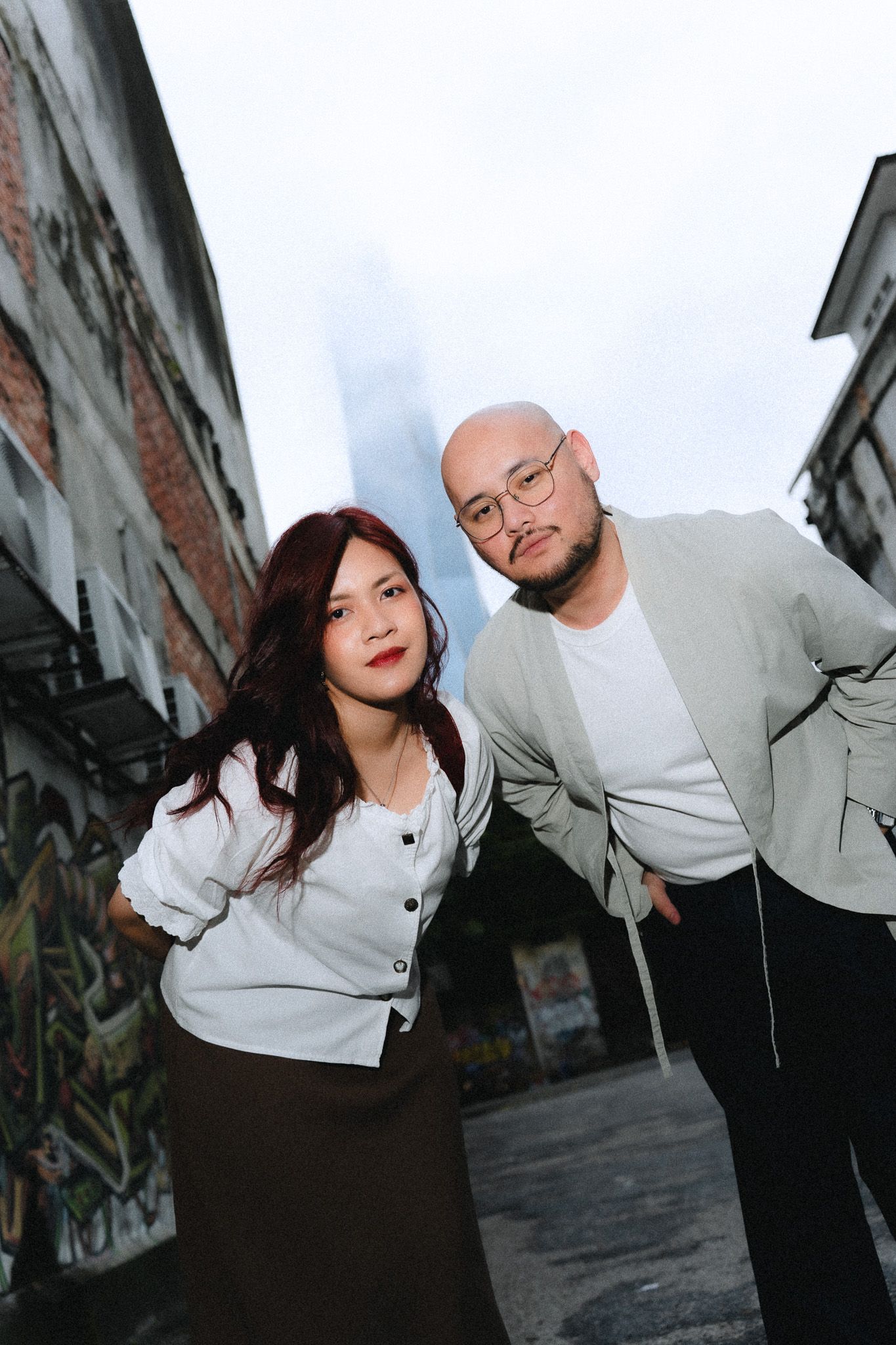 Two people pose confidently in an urban alley with graffiti walls and a skyscraper backdrop.