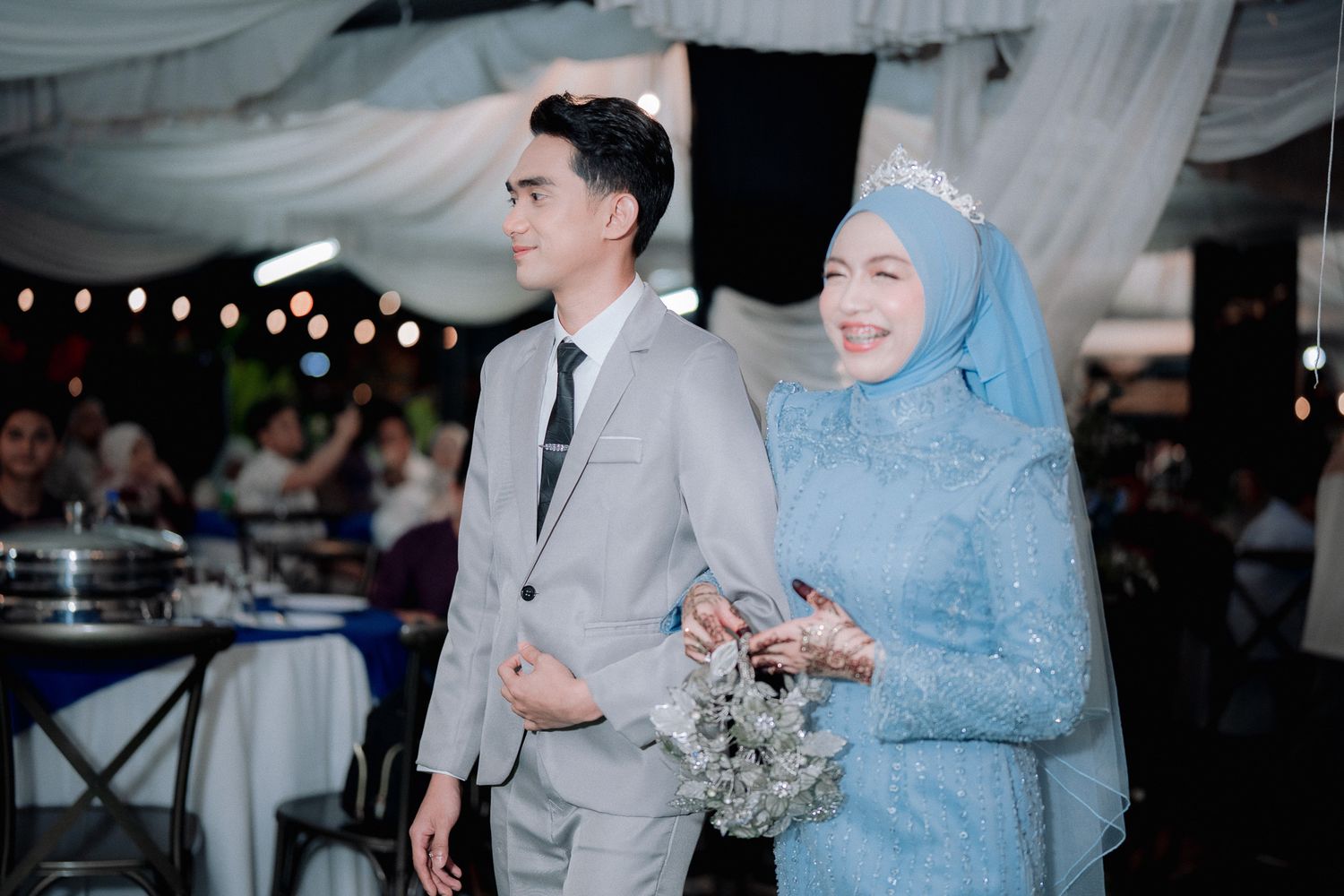 Muslim bride in blue hijab and groom in gray suit walking together at elegant wedding reception.