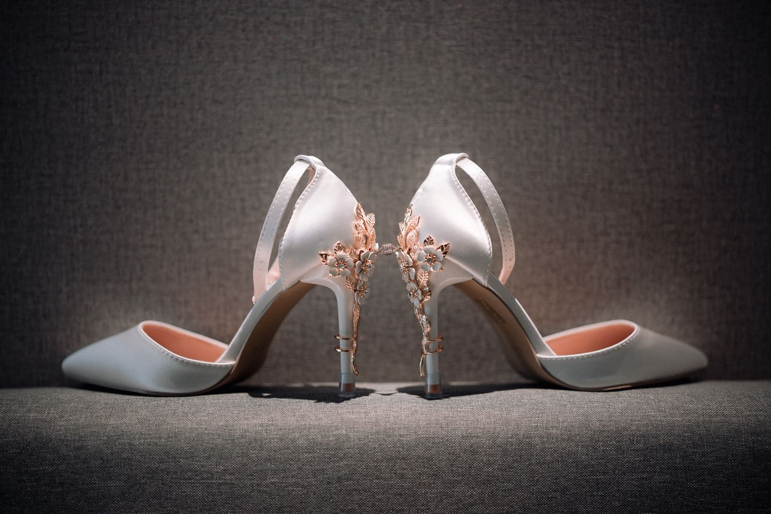 Elegant white satin bridal heels with delicate floral embellishments on the back, displayed on a gray fabric surface.