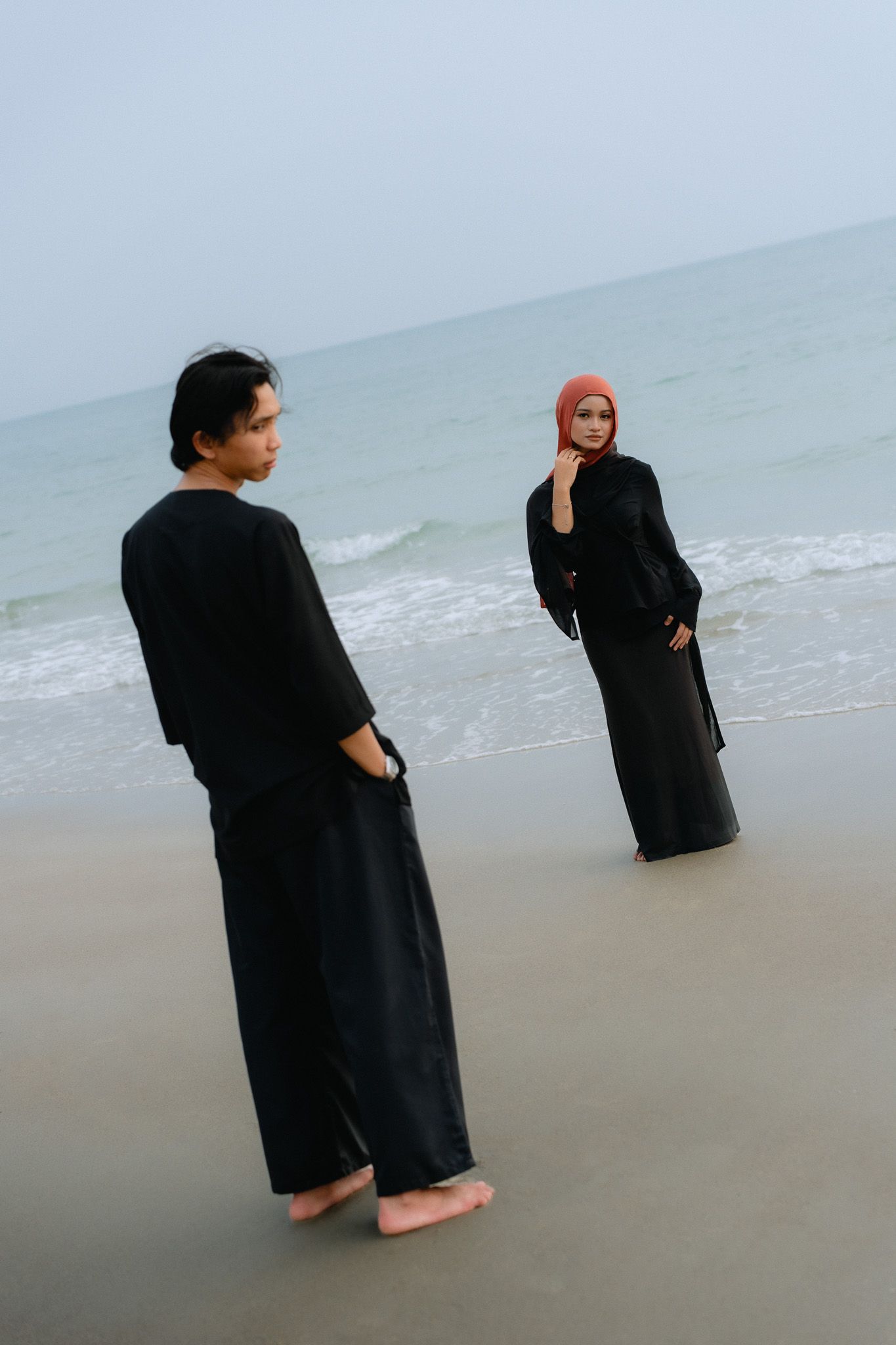 Couple in black outfits on beach, woman in hijab facing man near ocean waves.