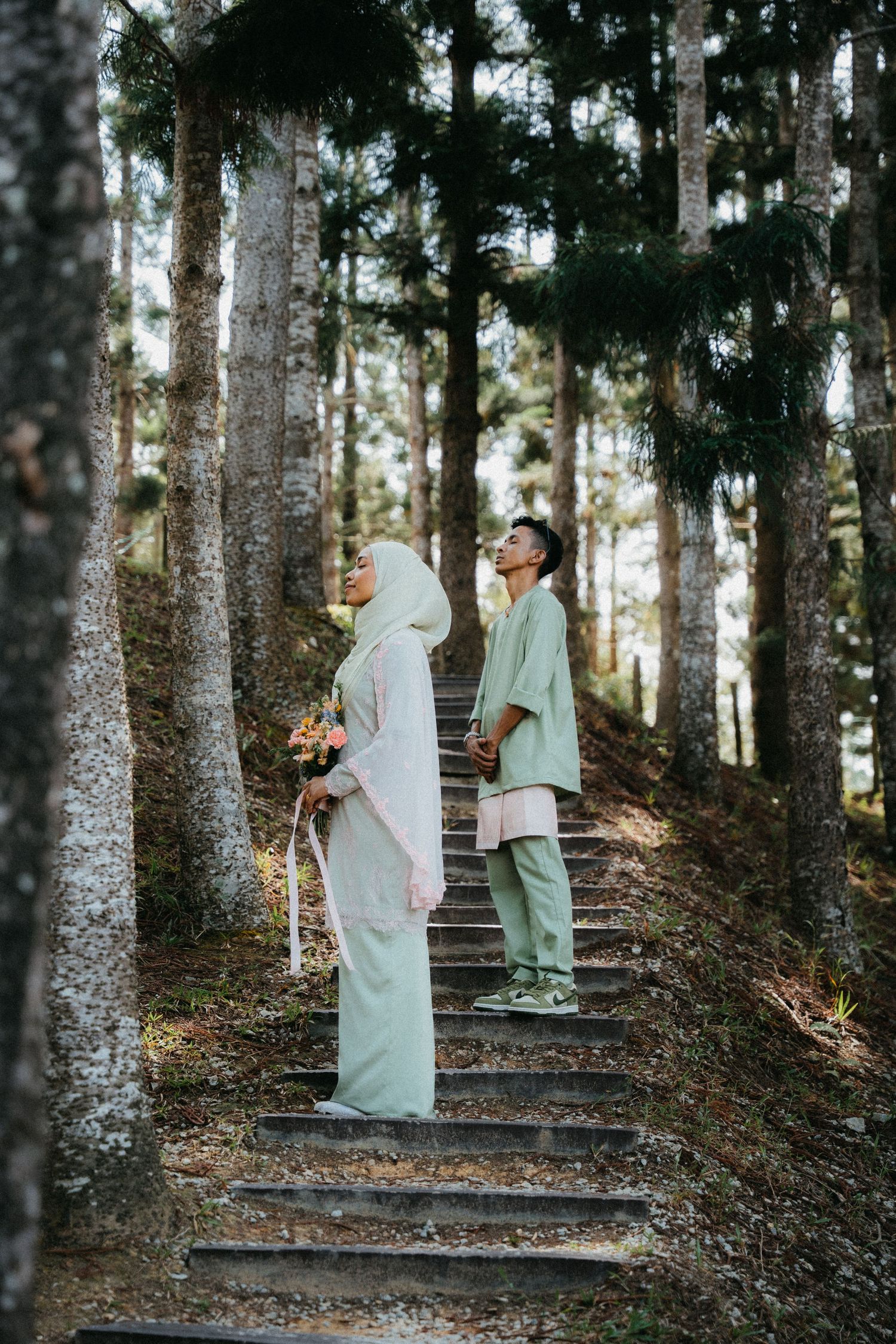 Two people standing on a rustic forest staircase wearing light colored clothing as sunlight filters through pine trees.
