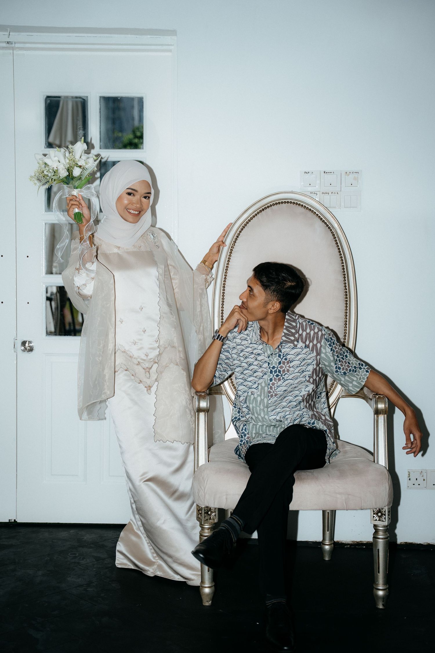 Two people lounging and interacting expressively on an elegant cream-colored chair against a white wall interior.
