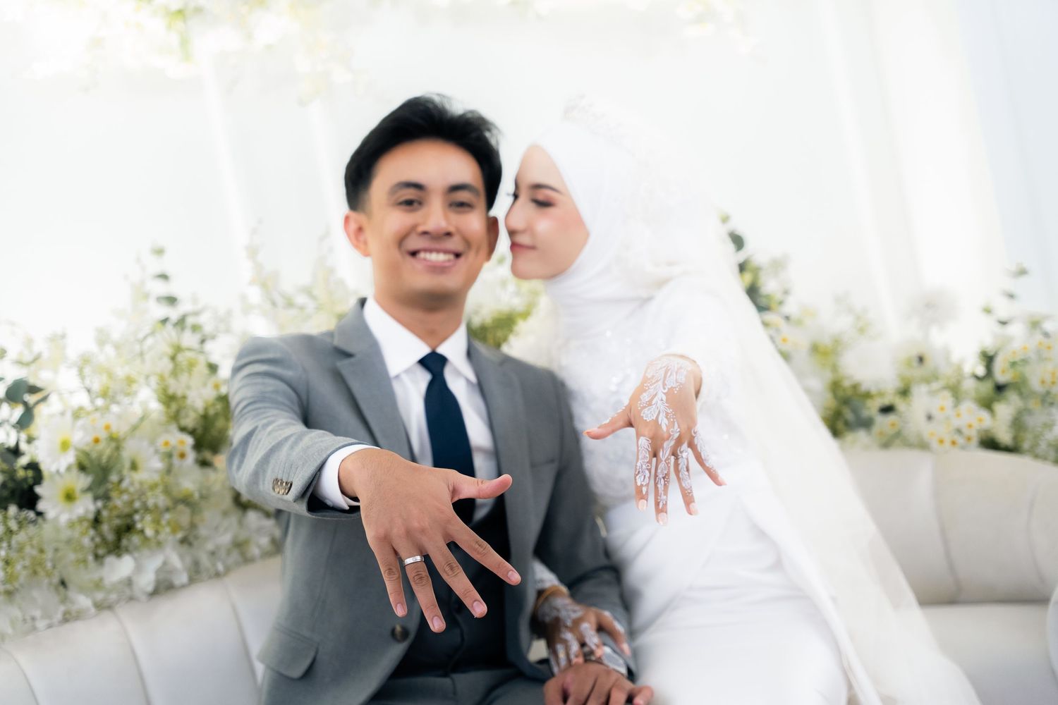 A happy newly married couple in formal wedding attire showing their rings against a white floral backdrop.
