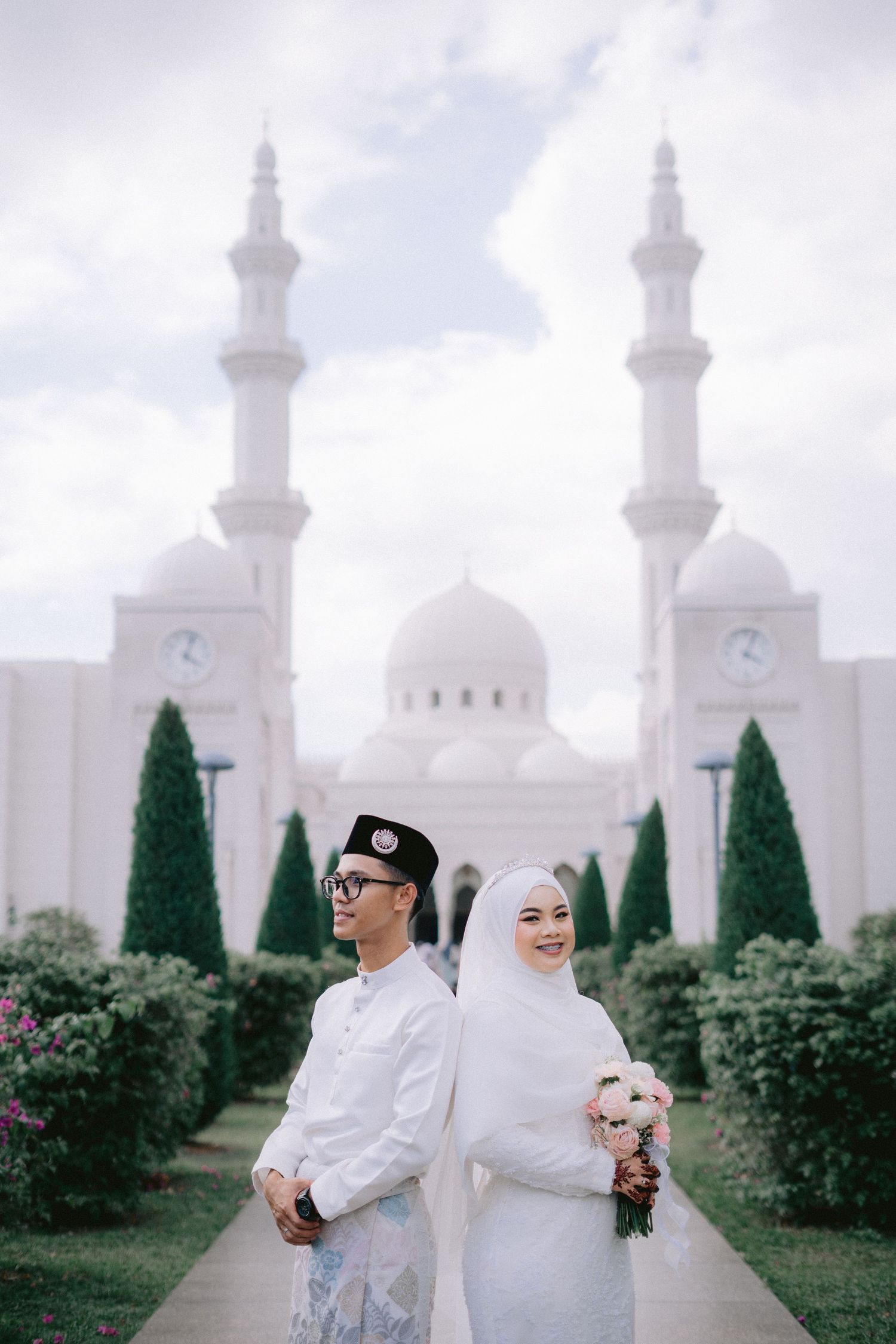 A couple in traditional white Muslim wedding attire poses in a garden with white minarets and cypress trees in the background.