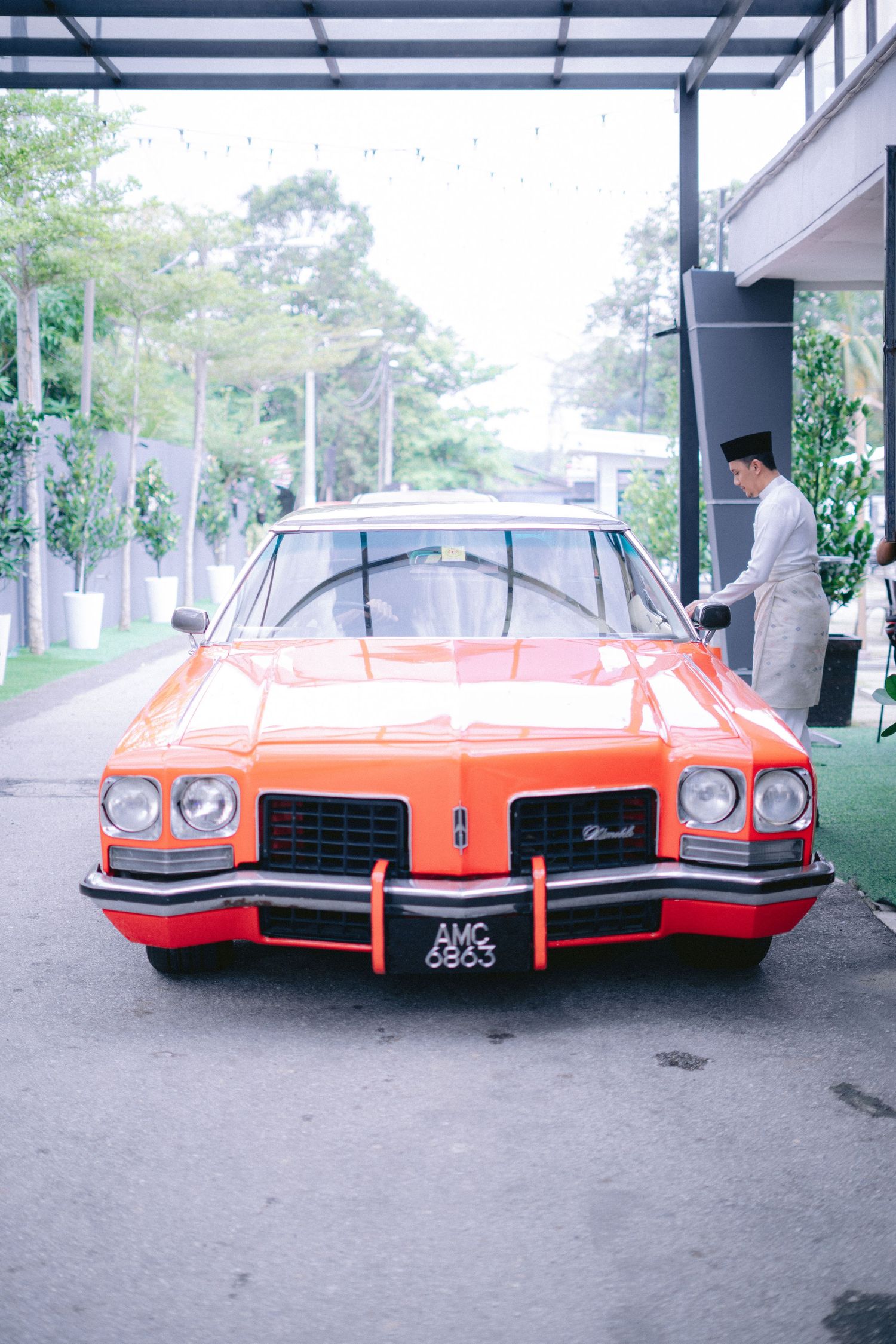 Classic orange Oldsmobile parked under a covered area with person standing beside it and greenery in background.