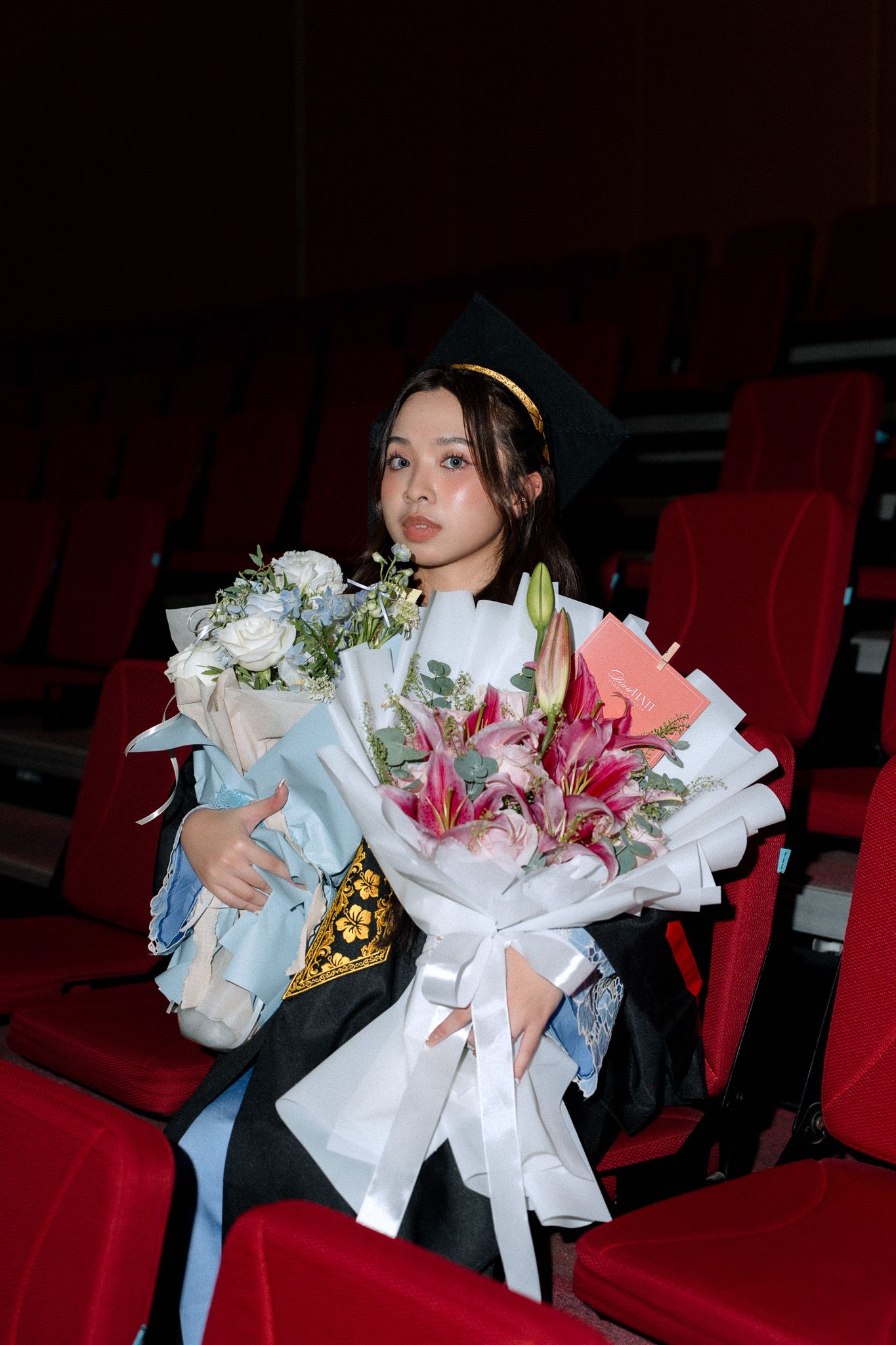 Young woman in graduation cap and gown holds colorful flower bouquets while seated in red auditorium chairs.