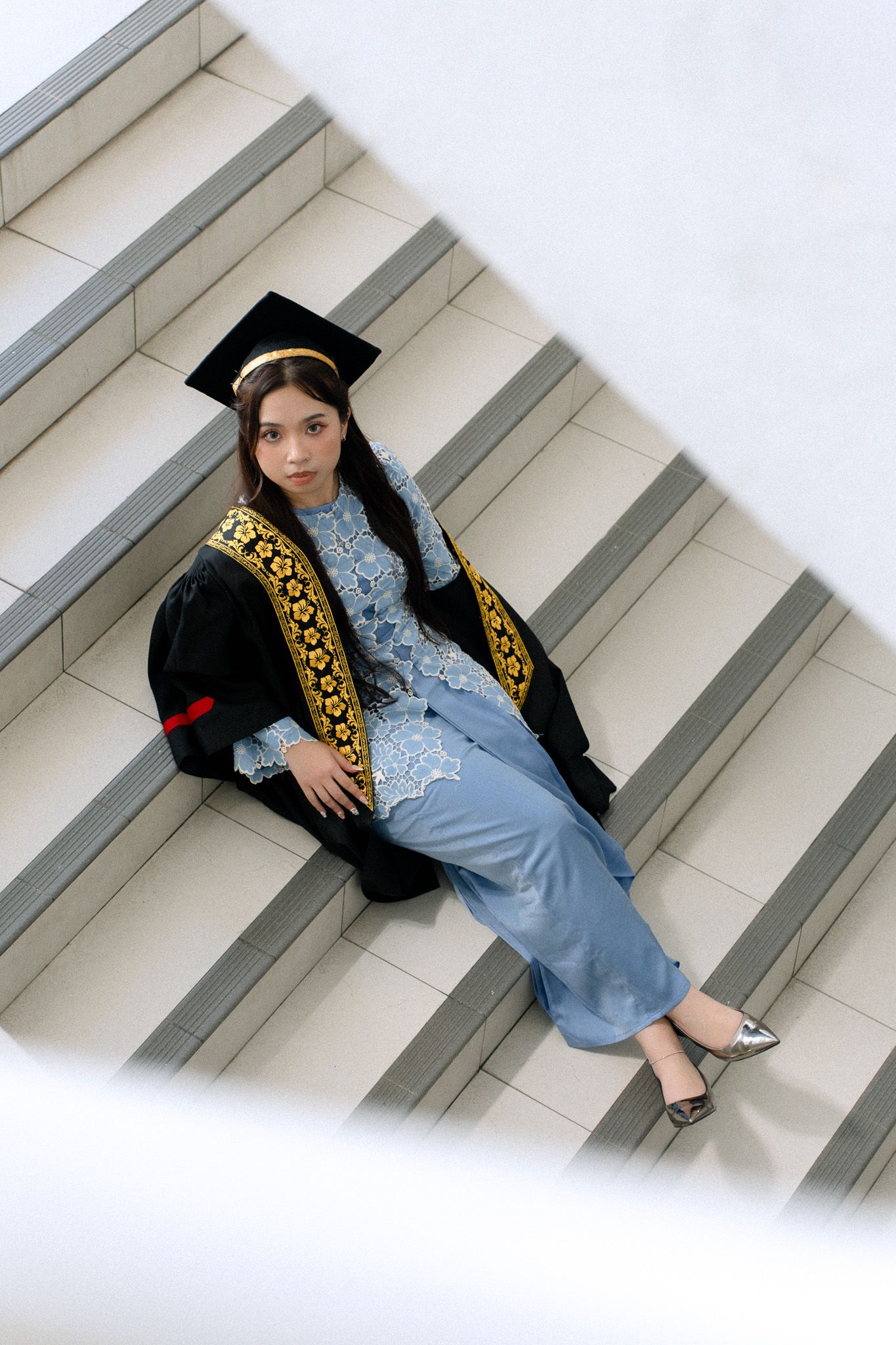 Young woman in graduation gown and cap sitting on white marble stairs, wearing blue traditional outfit.