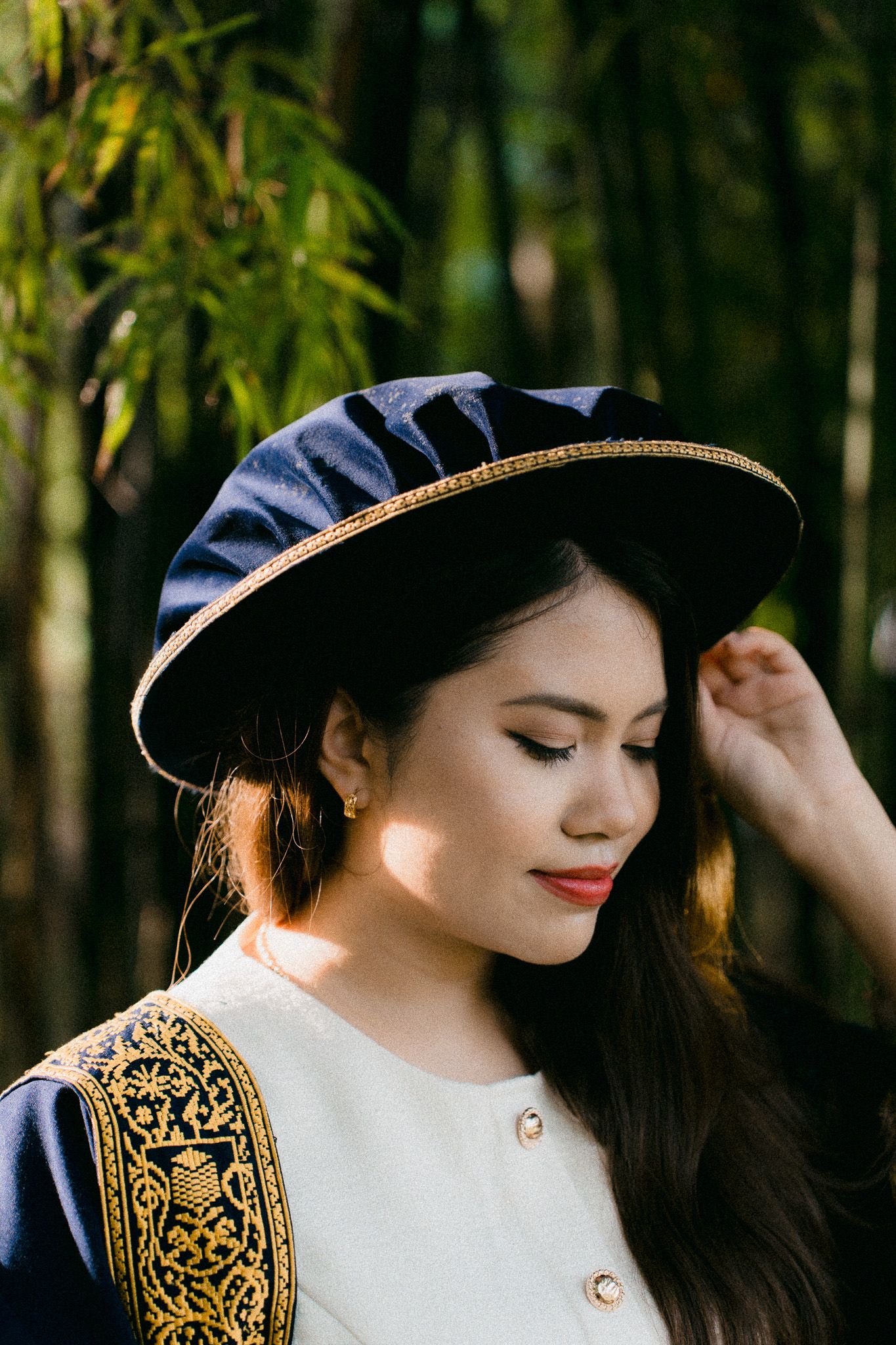 Young woman in traditional attire with ornate black and gold graduation hat poses outdoors among bamboo trees.