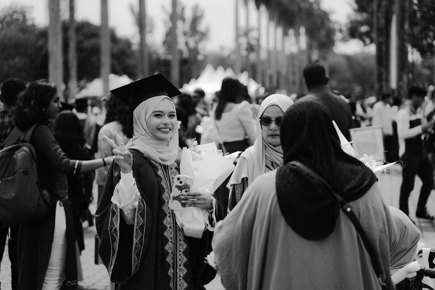 Black and white photo of a Muslim woman in graduation cap and gown smiling outdoors at a university ceremony.