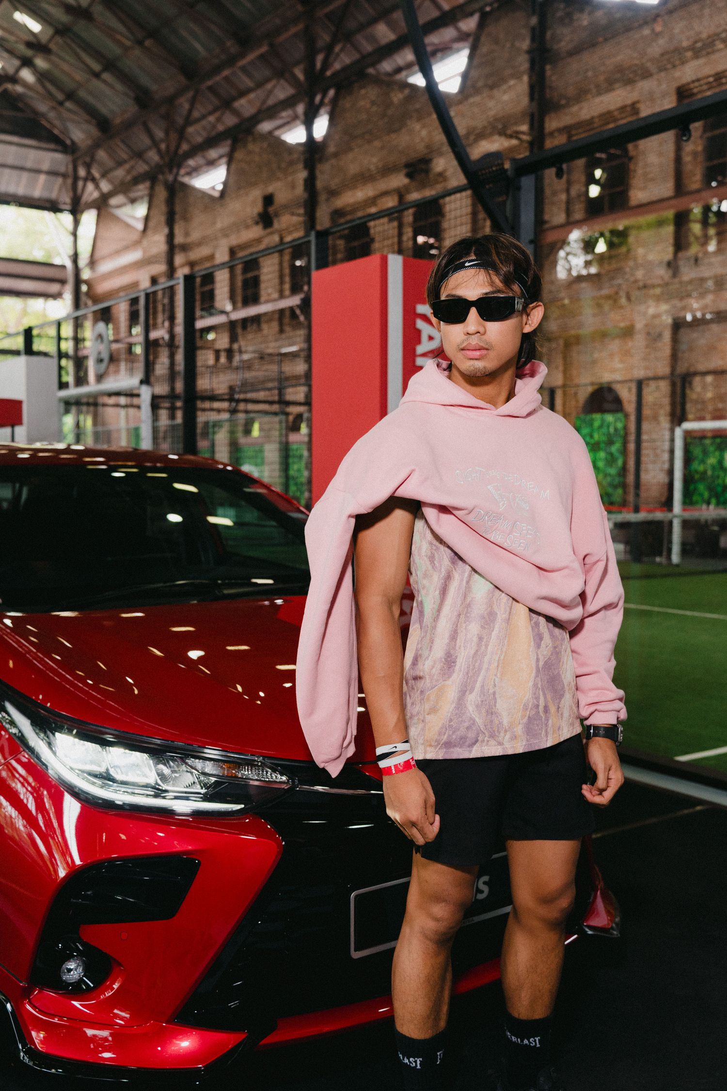 A person in a pink sweater and black shorts poses next to a red sports car in an indoor parking garage.