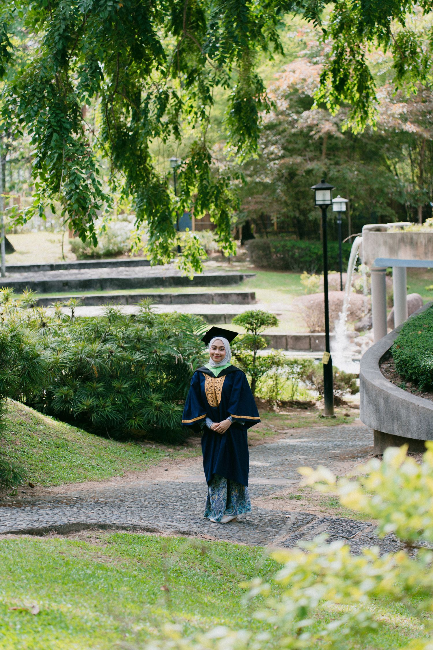 Graduate in academic regalia stands on a winding garden path beneath overhanging trees and lamp posts.
