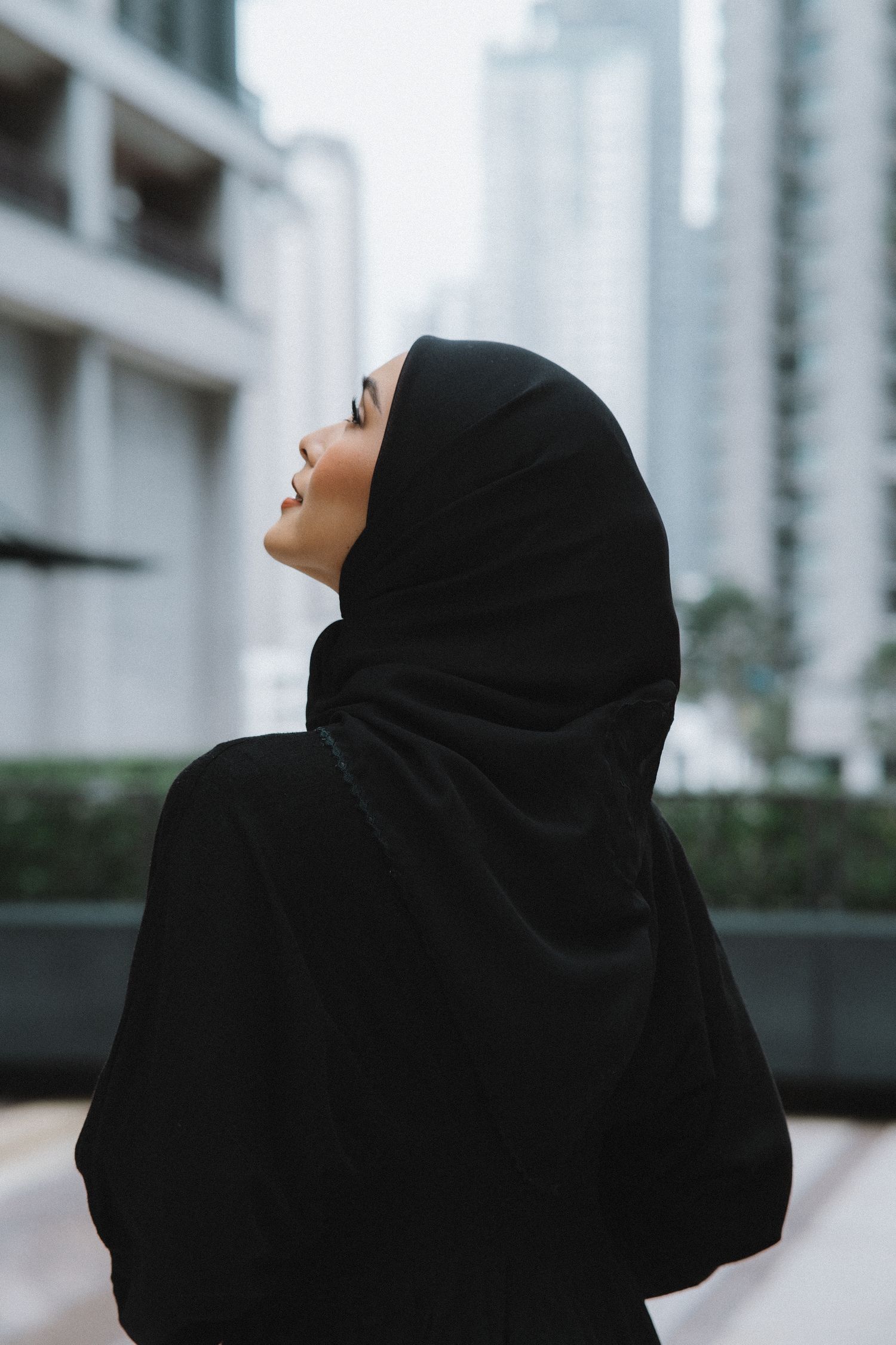Woman in black hijab gazes upward against urban backdrop of tall buildings.