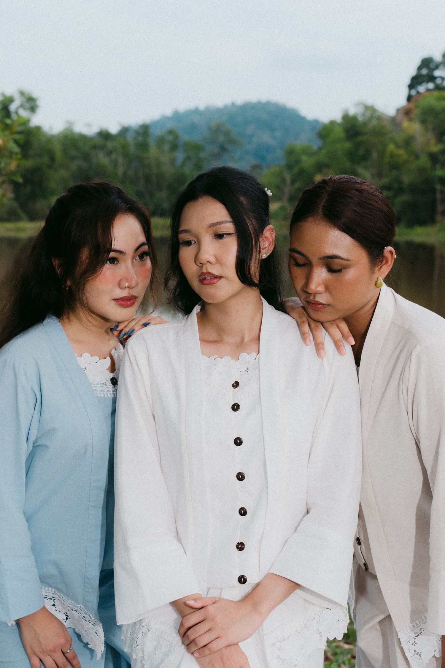 Three women in soft button-down shirts pose intimately together against a mountain backdrop.