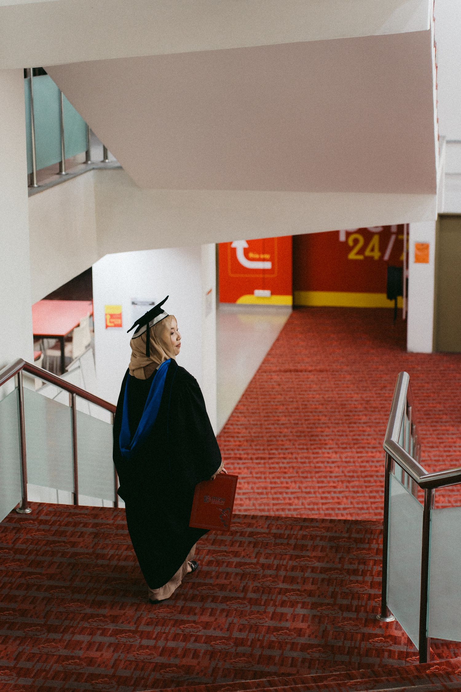 A graduate in cap and gown stands on red carpeted stairs in a modern indoor building hallway.