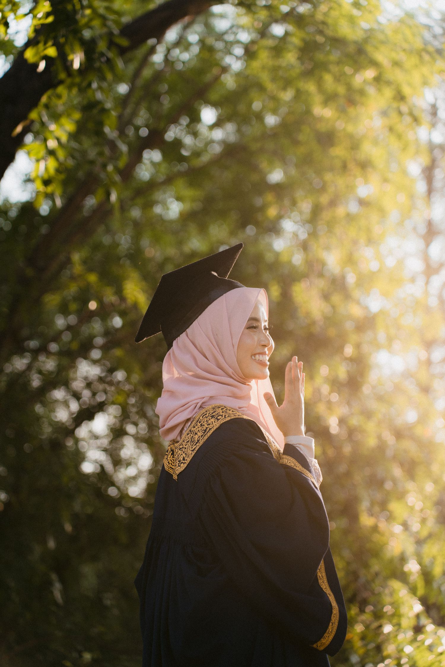 A graduate in black academic regalia and pink hijab smiles joyfully in golden sunlight during an outdoor celebration.