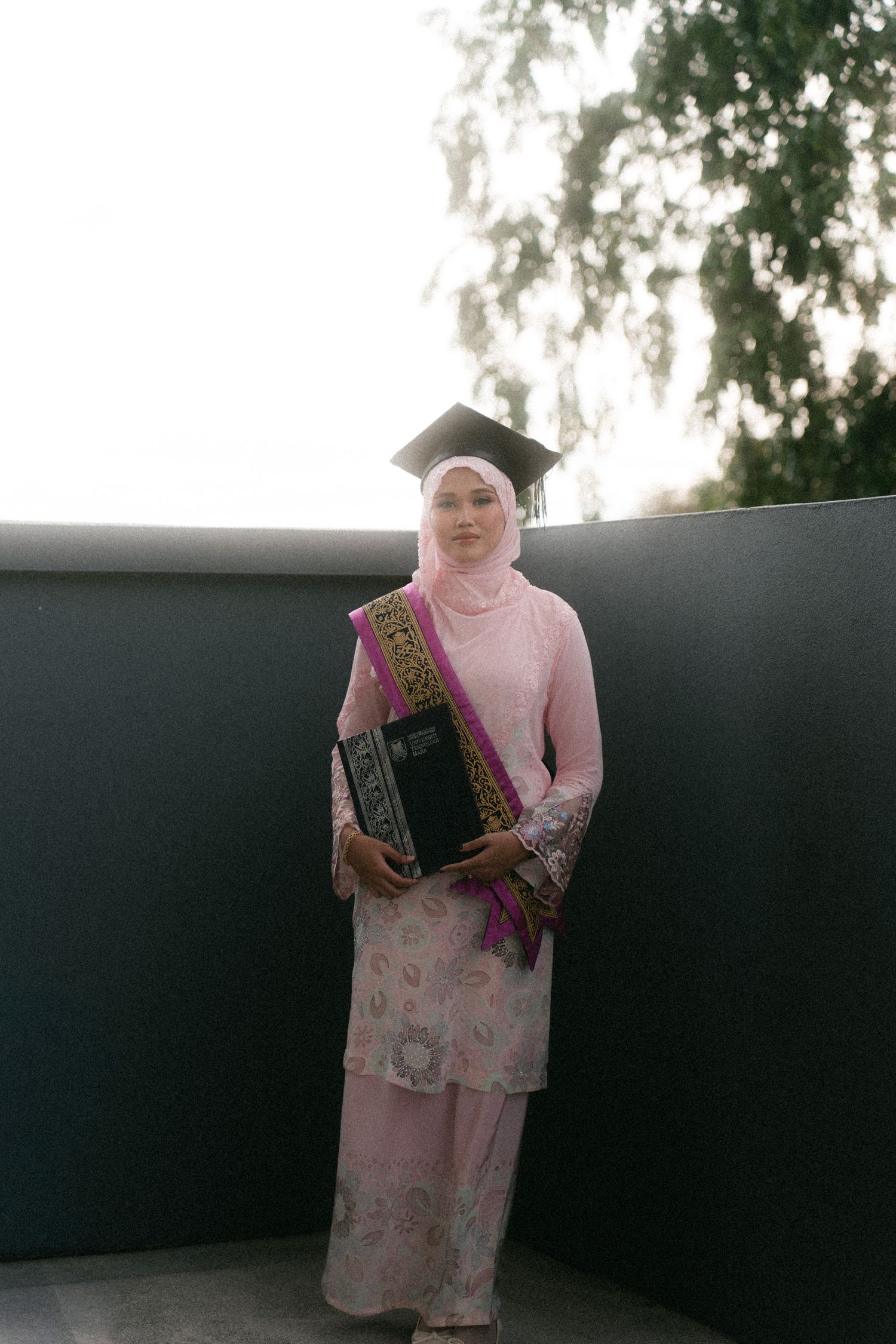A person in a pink dress and hijab holds a book while standing against a dark wall in multiple poses.