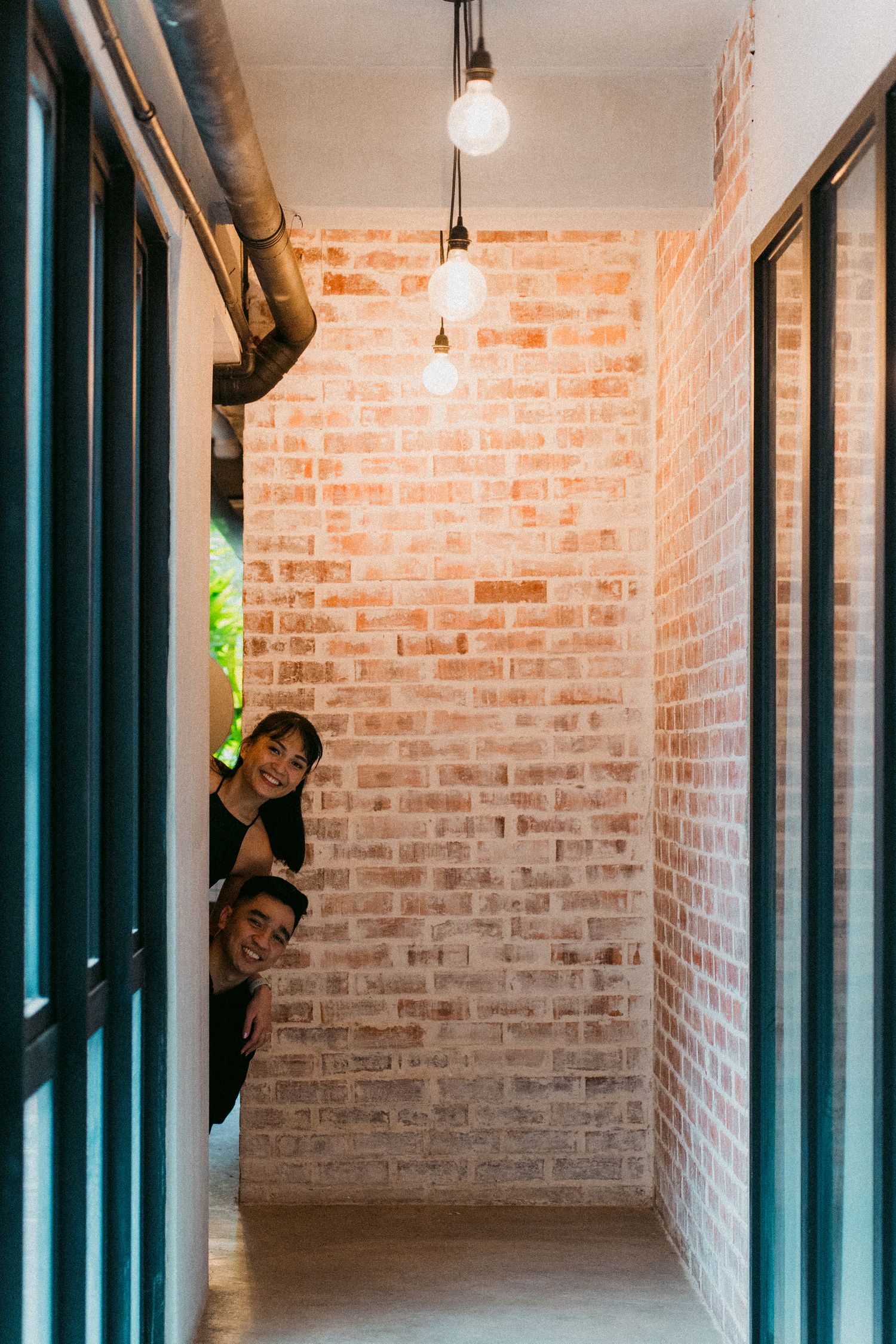 Playful couple peeking around corner of brick wall corridor with hanging light fixtures.