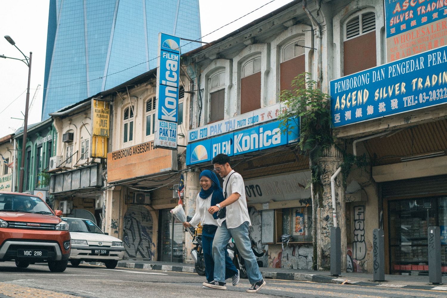 Two people walk past vintage shopfronts with Konica signage on an urban street.