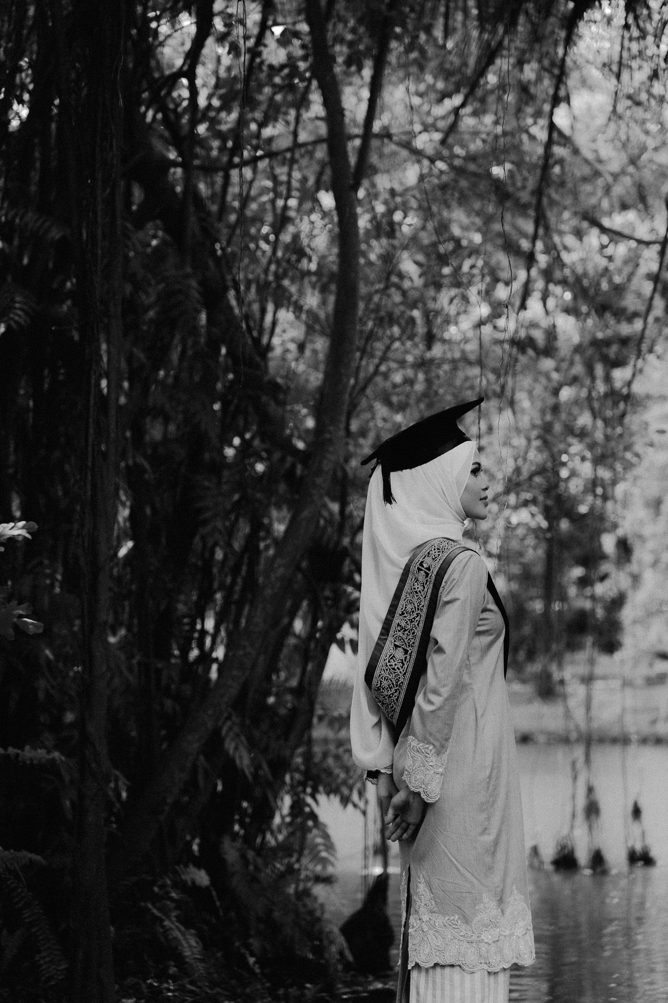 Black and white photo of a Muslim graduate in hijab and mortarboard cap standing among trees near a lake.