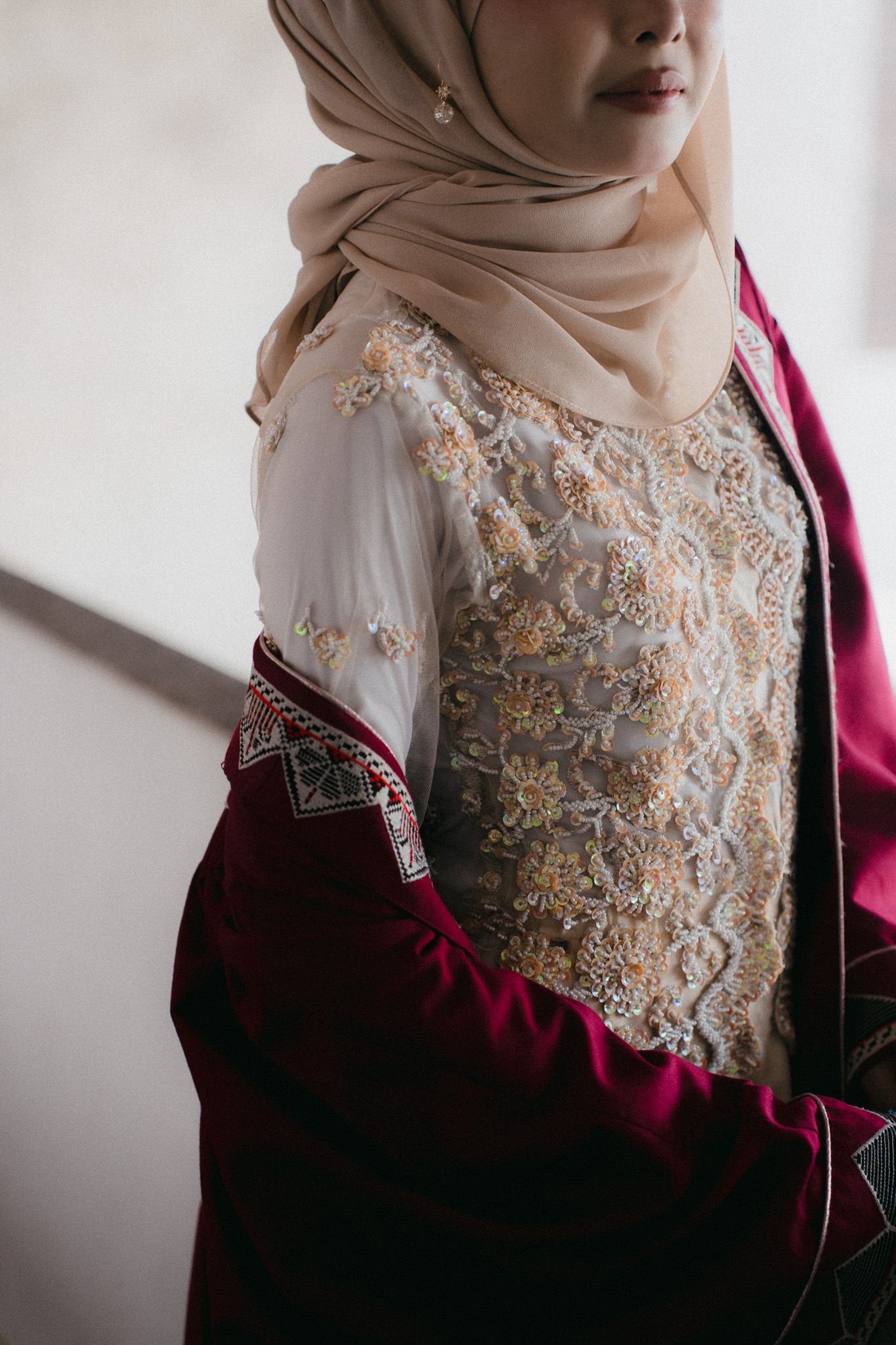 A woman in a burgundy abaya with intricate gold embroidered lace overlay and beige hijab with pearl earring.