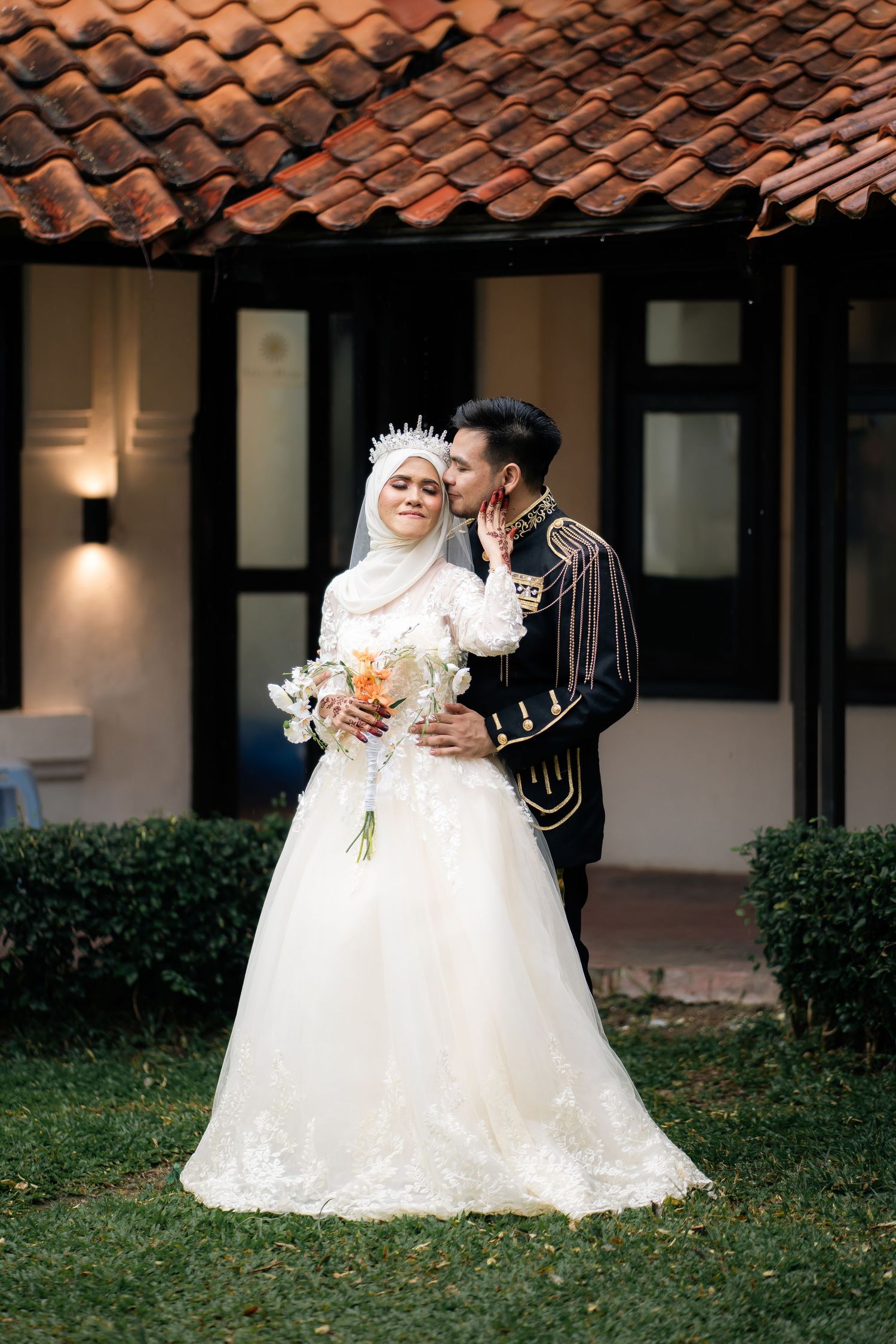 Beautiful outdoor wedding photo featuring a couple in traditional attire with white dress and hijab against terracotta roofed building.