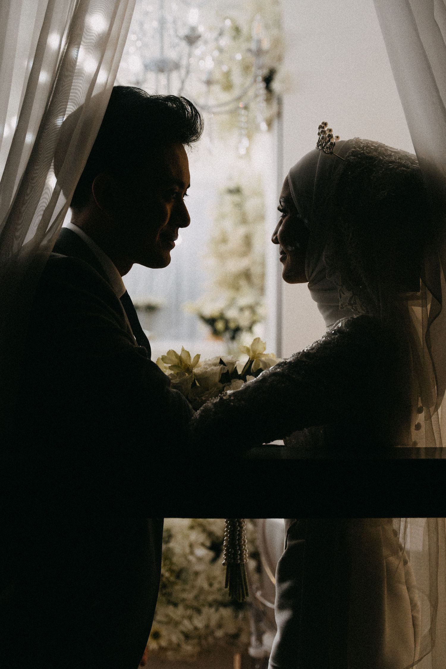 A silhouetted couple in wedding attire share an intimate moment by a sunlit window with sheer curtains.