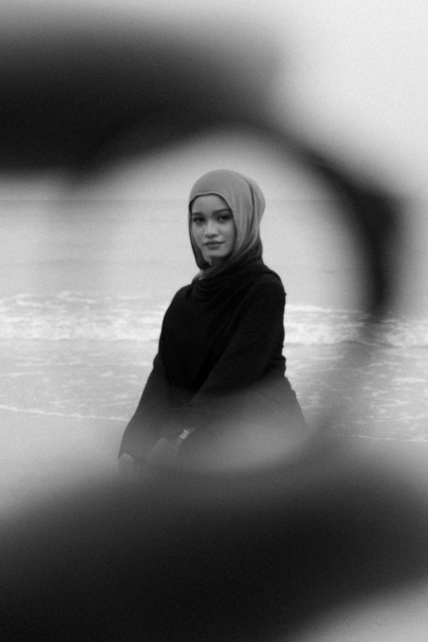 Black and white portrait of a young woman wearing a hijab sitting near the ocean, photographed through a blurred foreground frame.