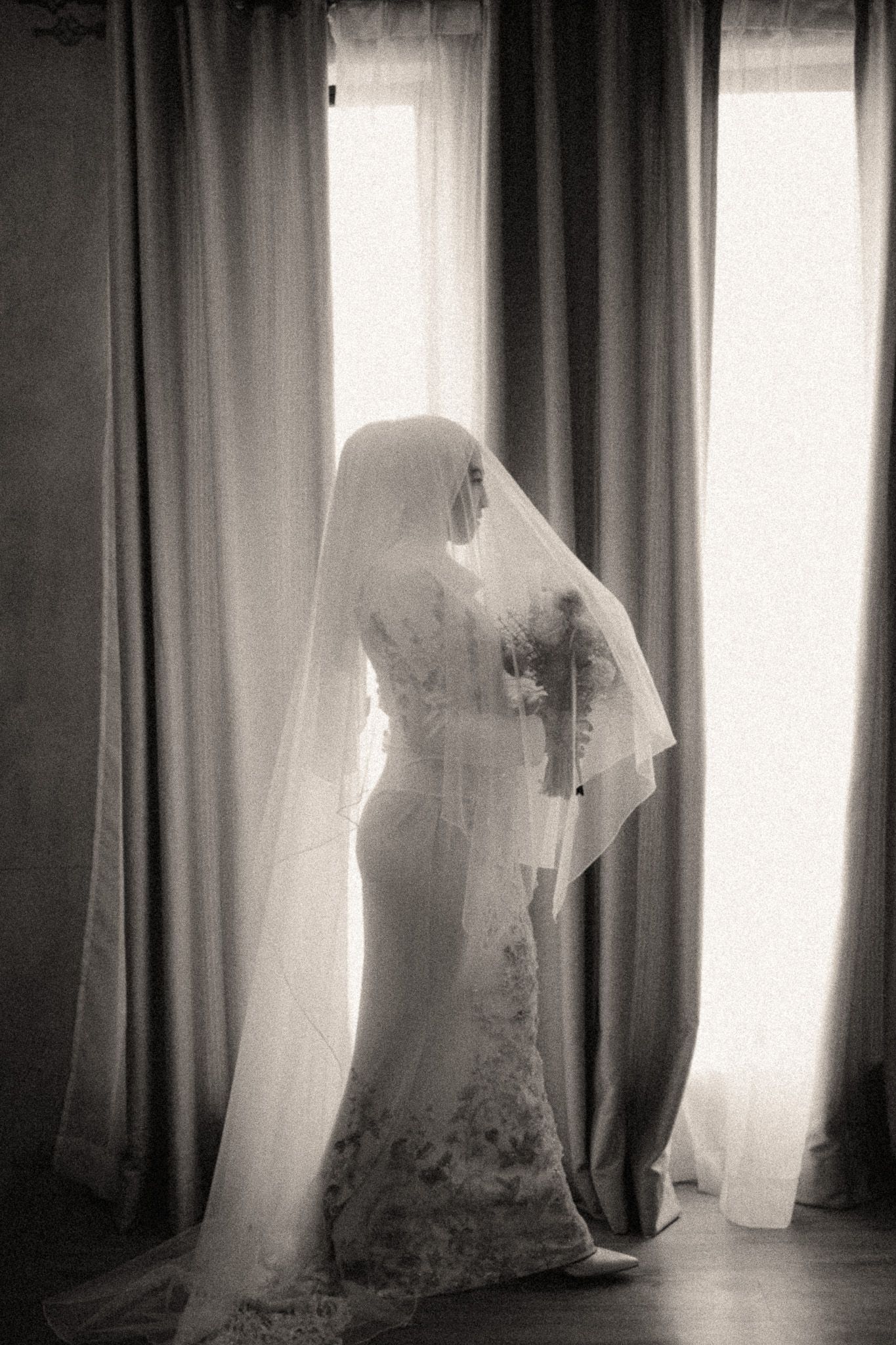 Black and white photo of a bride in lace gown and veil standing by a sunlit window on her wedding day.