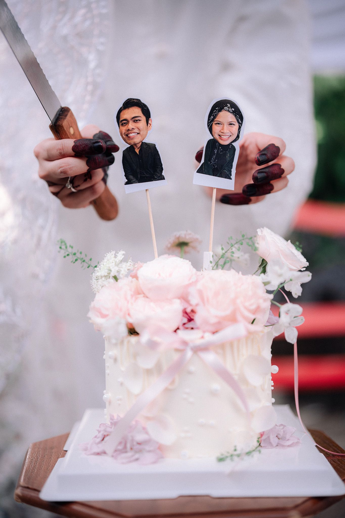 White wedding cake with pink flowers and custom couple face toppers on sticks, being cut with a knife.