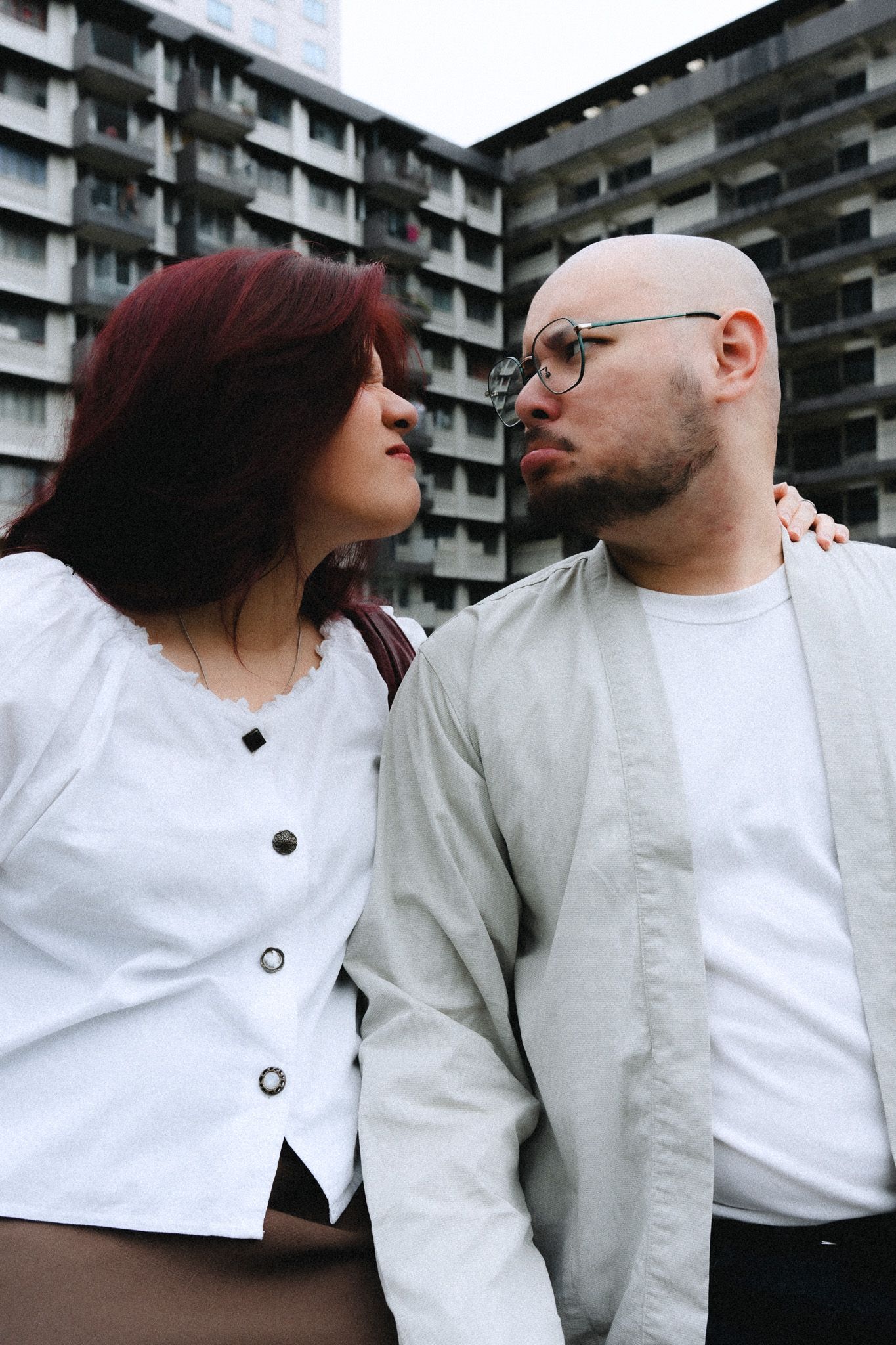 A bald man with glasses and a woman with red hair share a tender moment outdoors, with a urban building in the background.