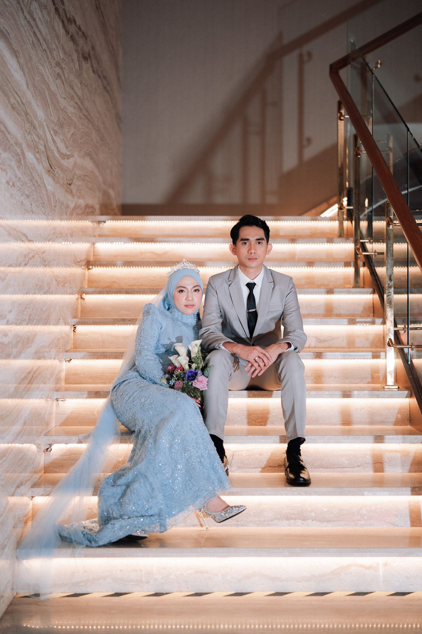Couple in formal attire sitting on illuminated marble stairs, bride in blue hijab holding bouquet.