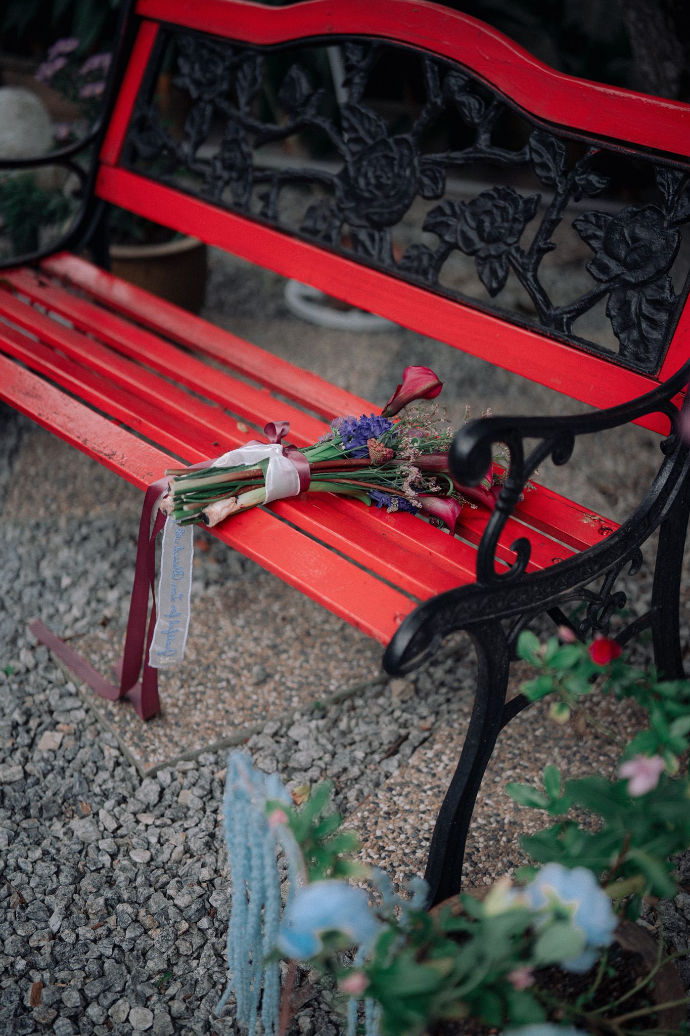 A vibrant red garden bench with ornate black ironwork holds a small wildflower bouquet tied with ribbon, set on gravel.