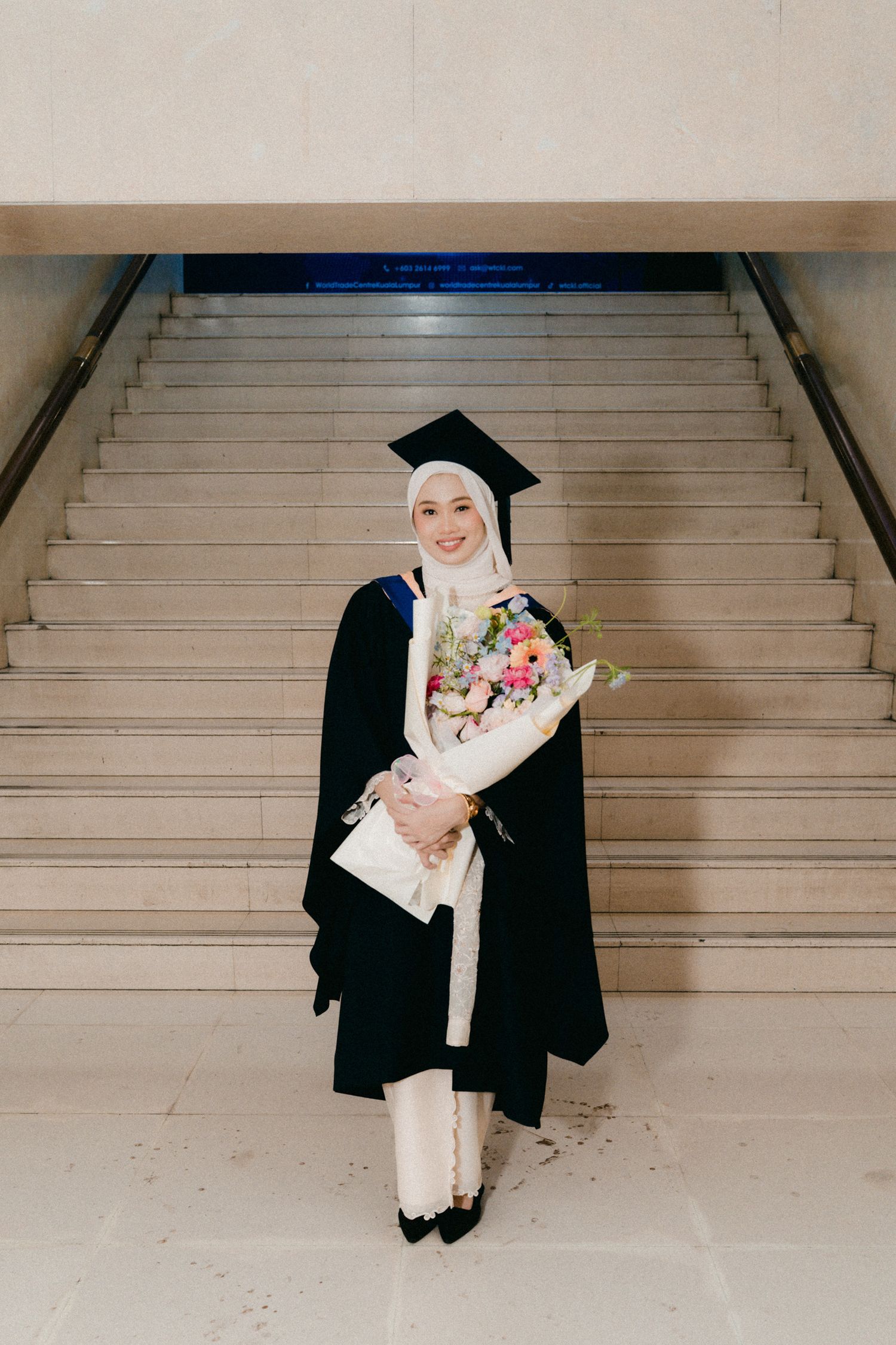 A graduate in black cap and gown holding a floral bouquet stands on stairs wearing a white hijab and formal attire.