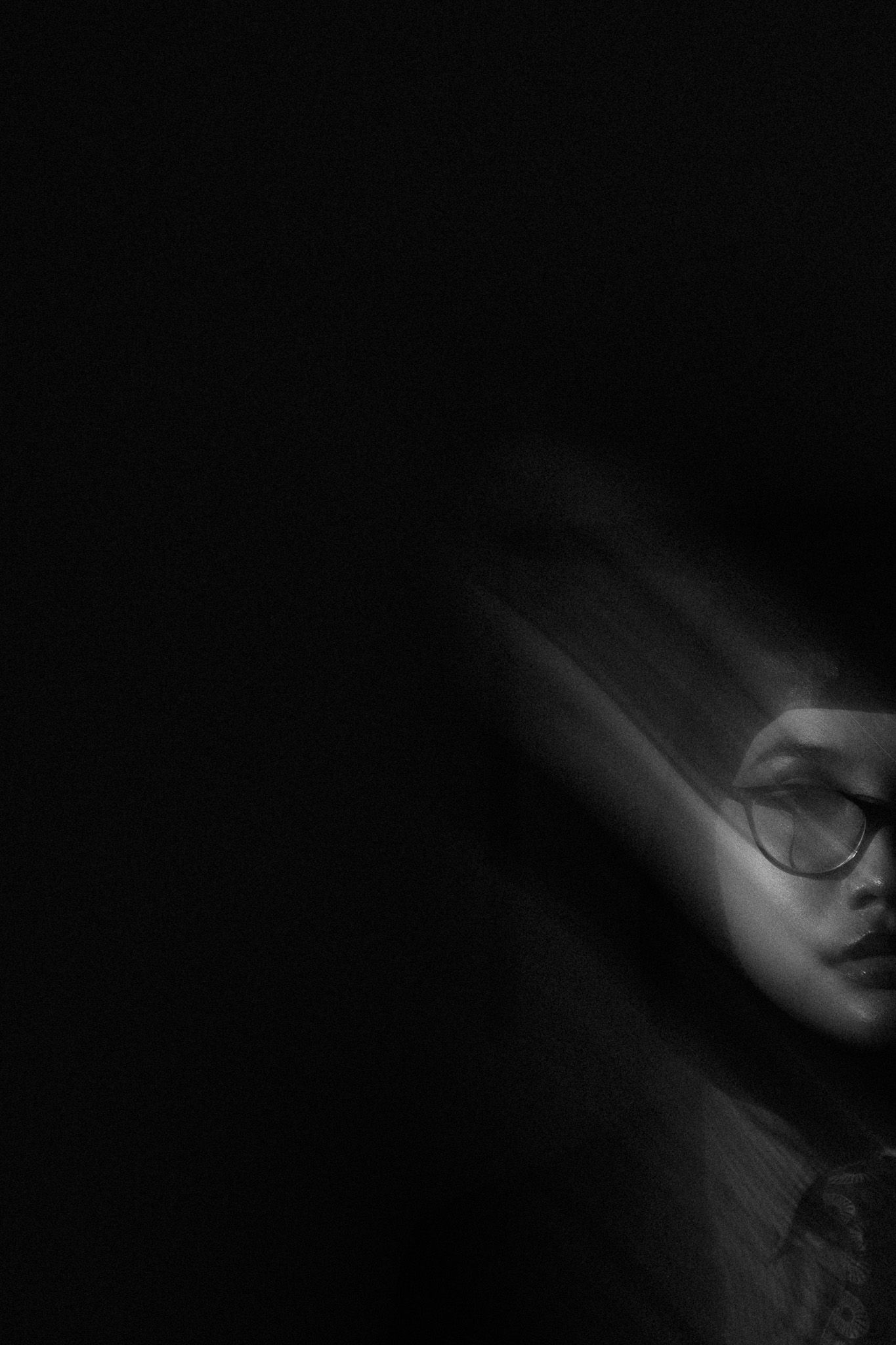 Blurred black and white portrait of a woman wearing round glasses emerging from darkness.