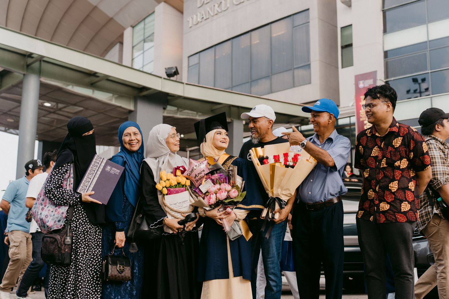 A group of people holding graduation bouquets and gifts gather outside a modern building during a celebration.