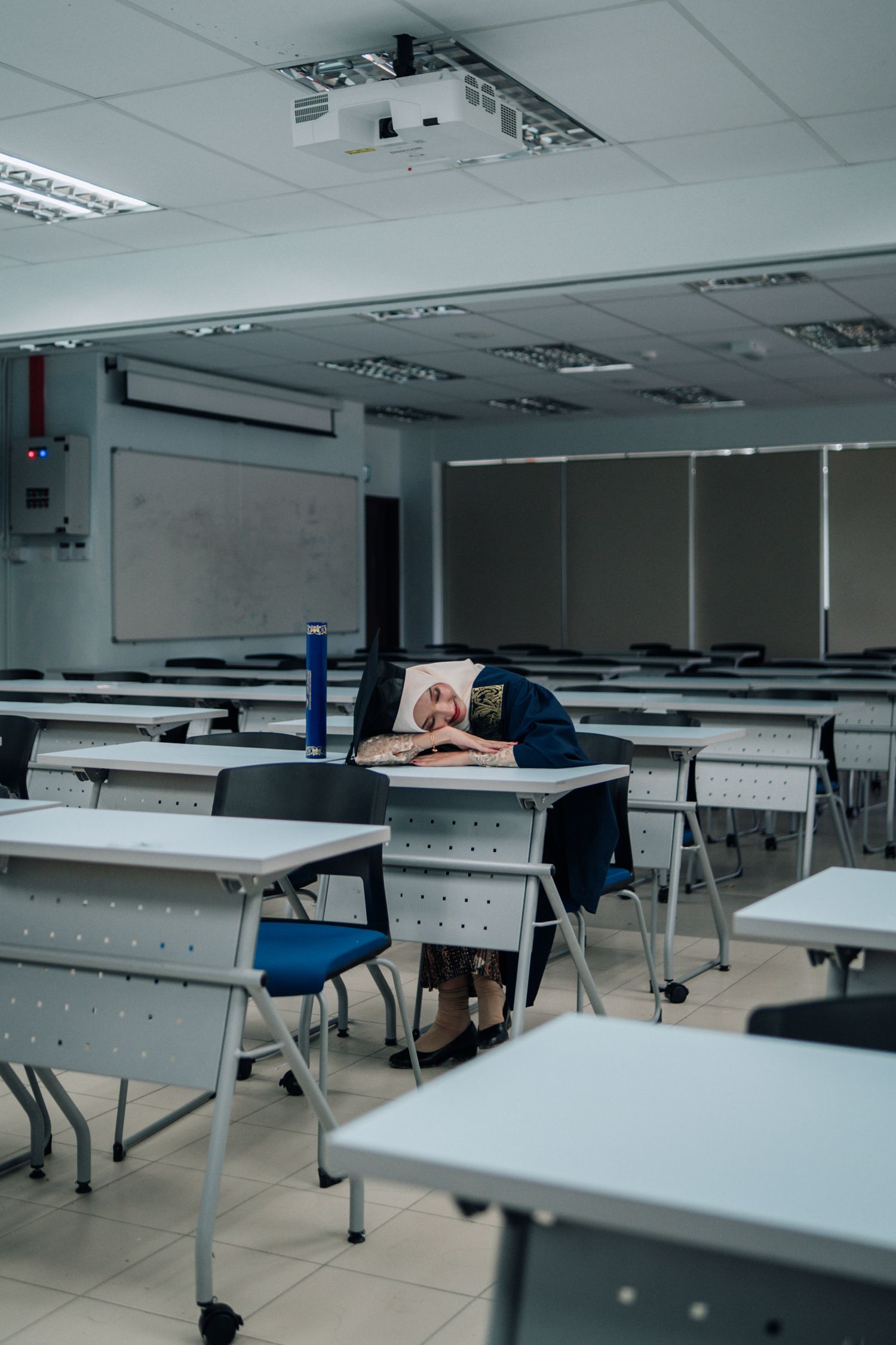 Student rests head on desk in empty classroom with fluorescent lighting and projector mounted on ceiling.