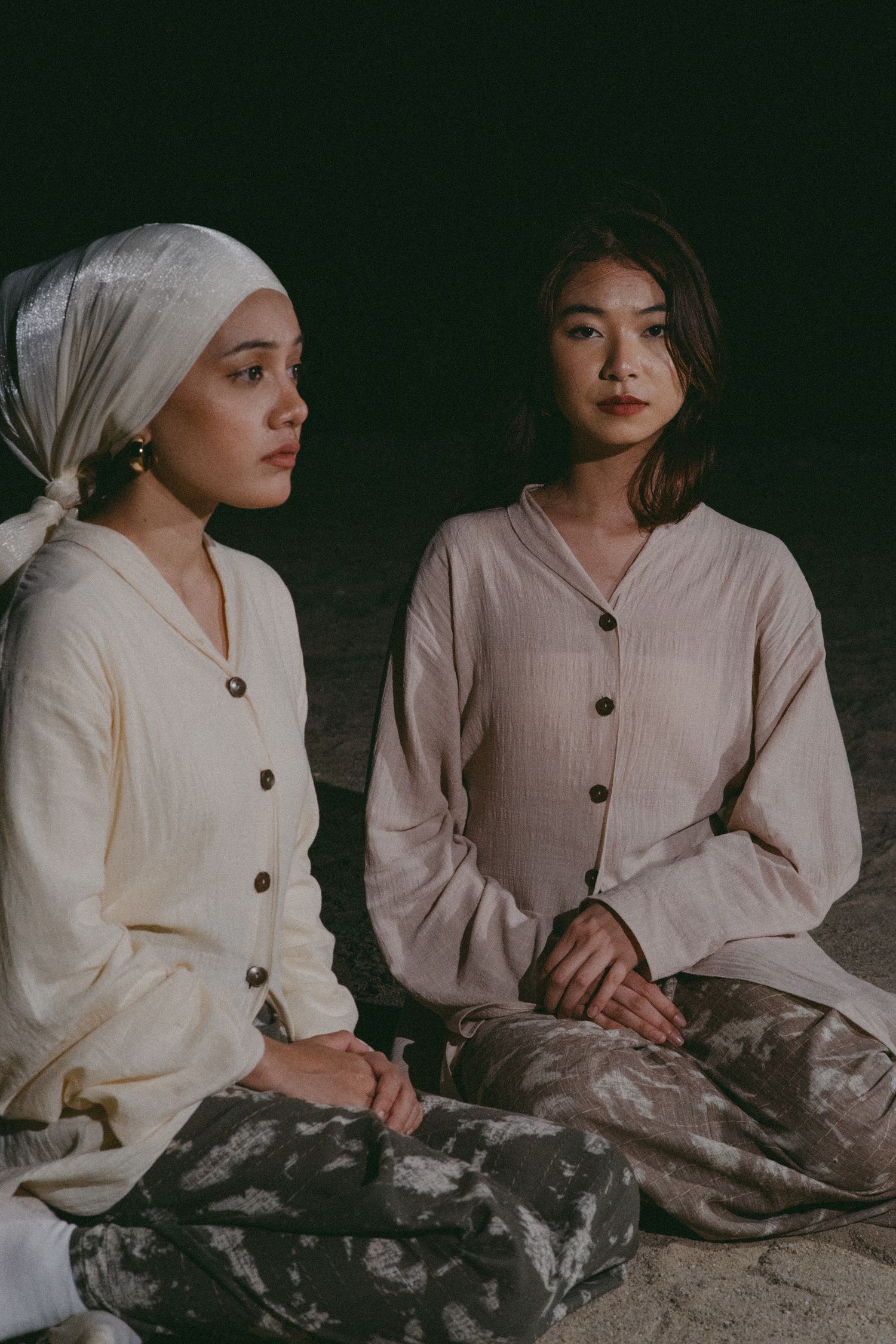 Two people in light colored clothing sitting together against a dark background.