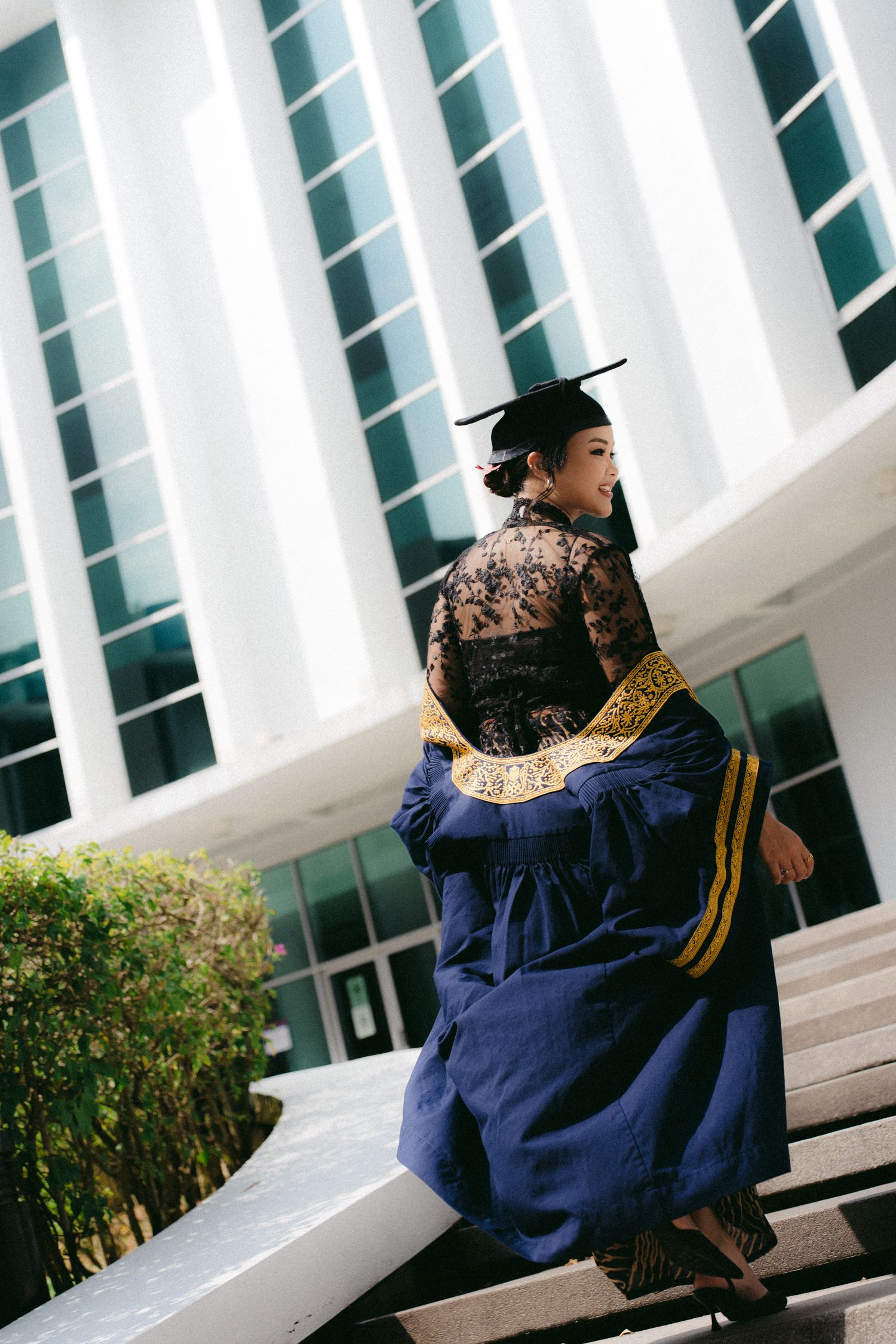 Graduate in navy blue cap and gown sits on steps of modern building with architectural white columns.