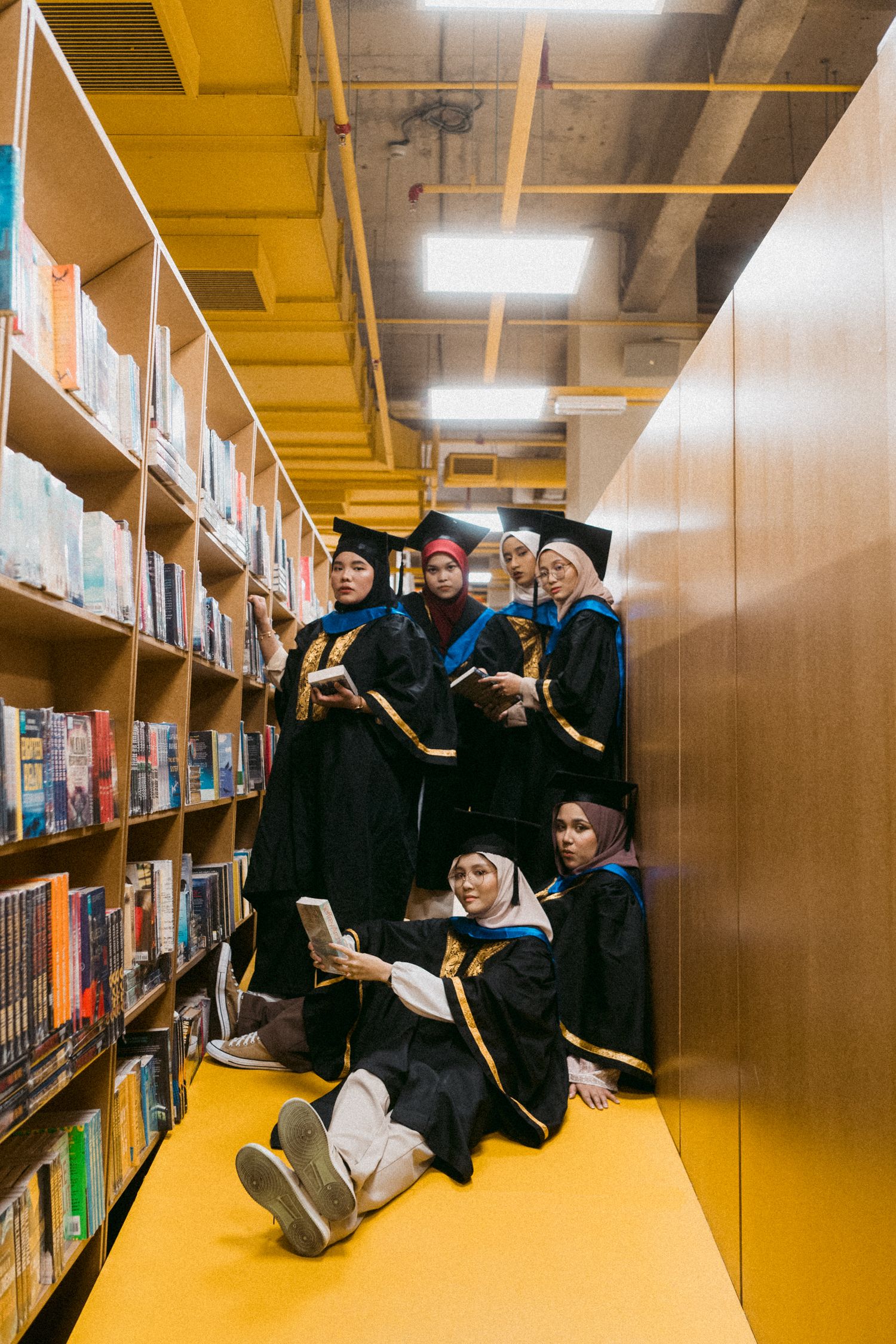 Graduates pose playfully between library bookshelves on yellow floor.