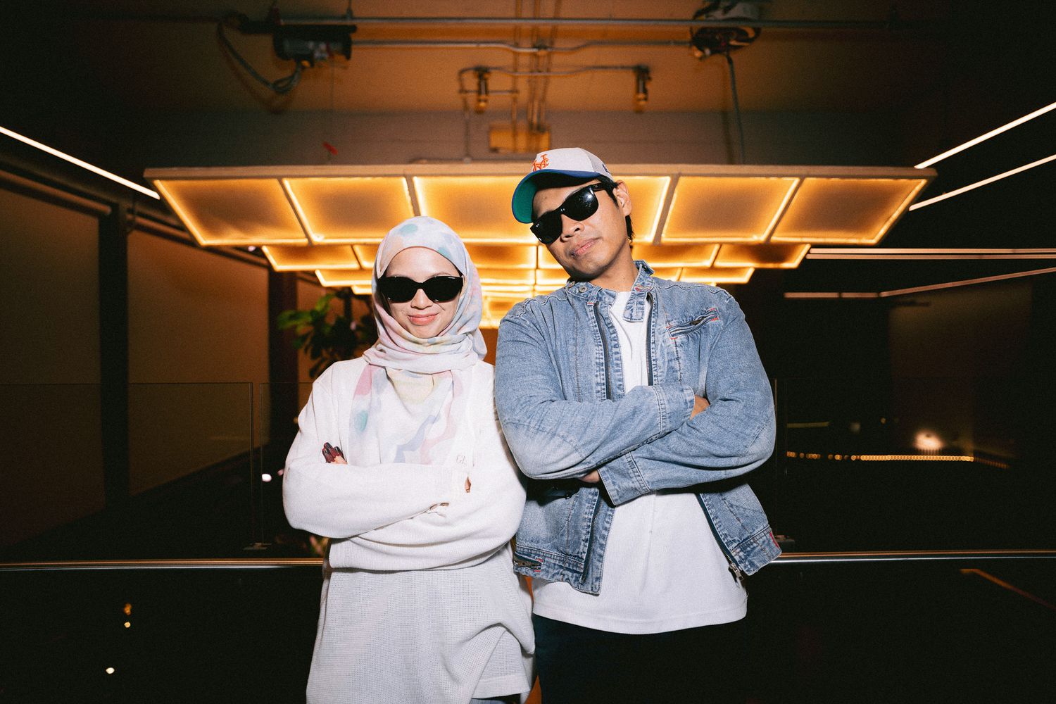 Two stylish individuals in sunglasses pose confidently with crossed arms under warm golden lights.
