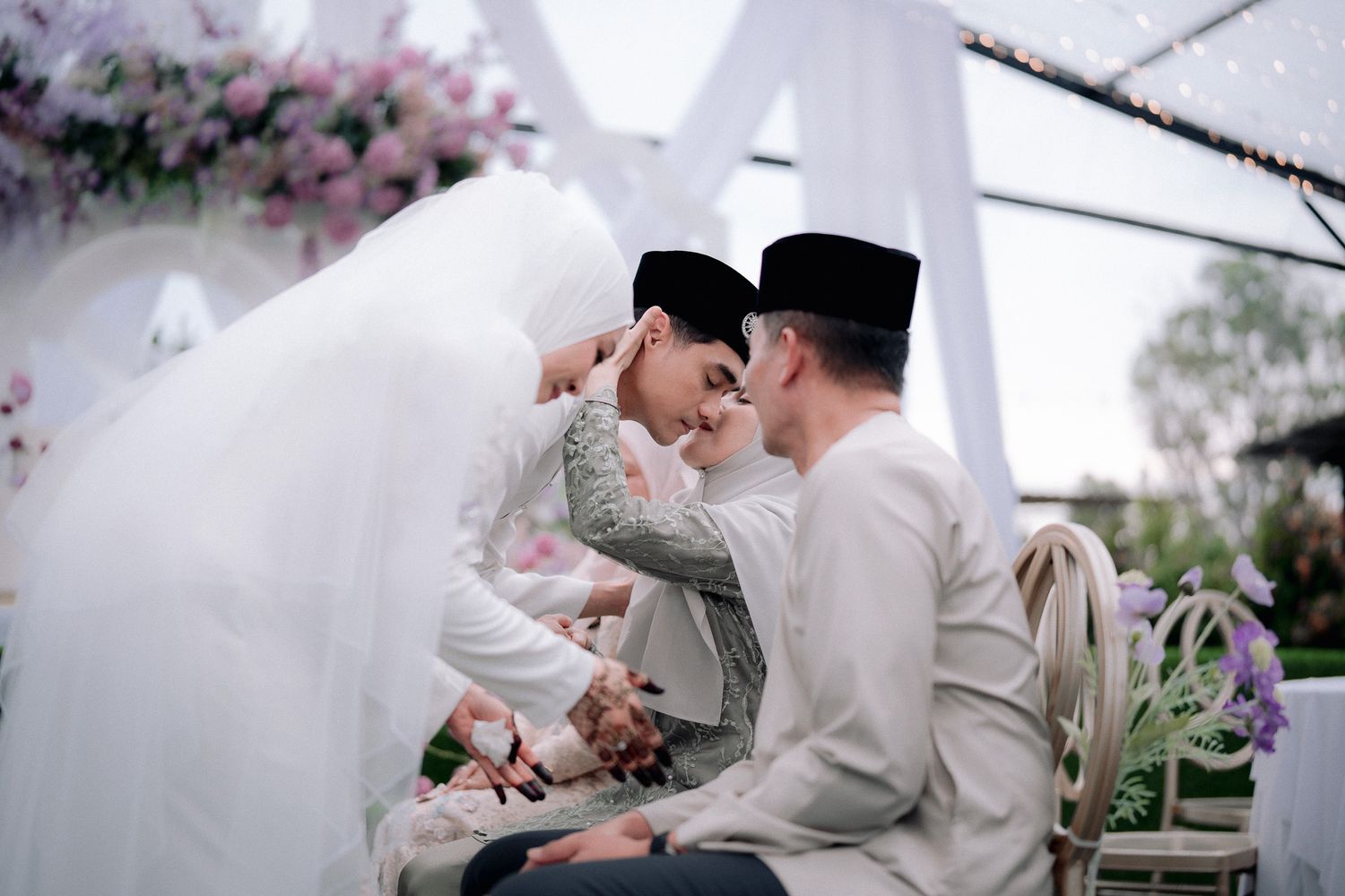 A Muslim couple in traditional white attire share an intimate moment at their wedding ceremony adorned with floral decorations.