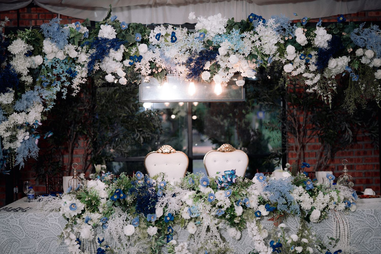 Elegant wedding sweetheart table adorned with blue and white floral arrangements under a floral arch with twinkling lights.
