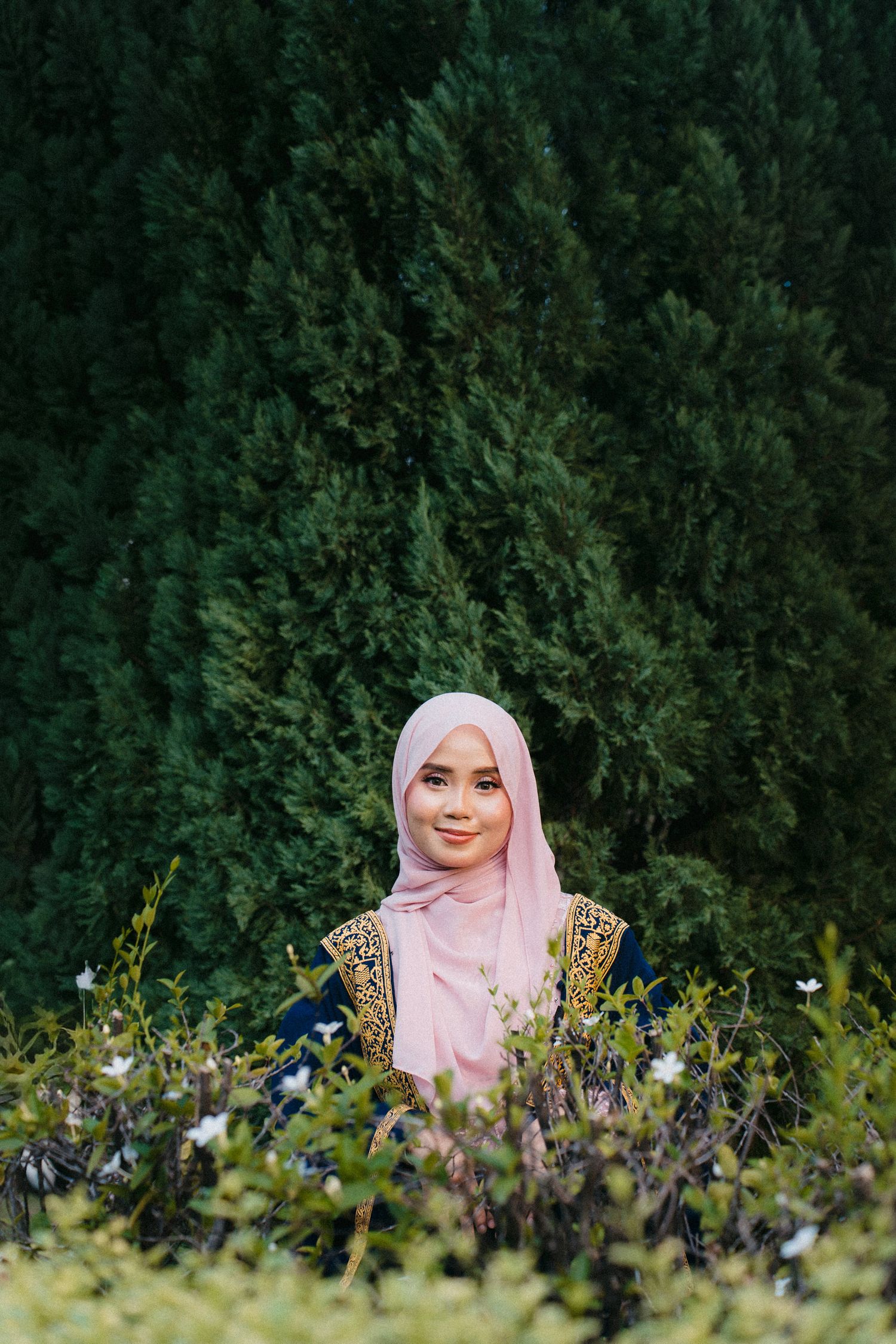 A person wearing a pink hijab smiles while sitting among wildflowers and greenery in a garden setting.