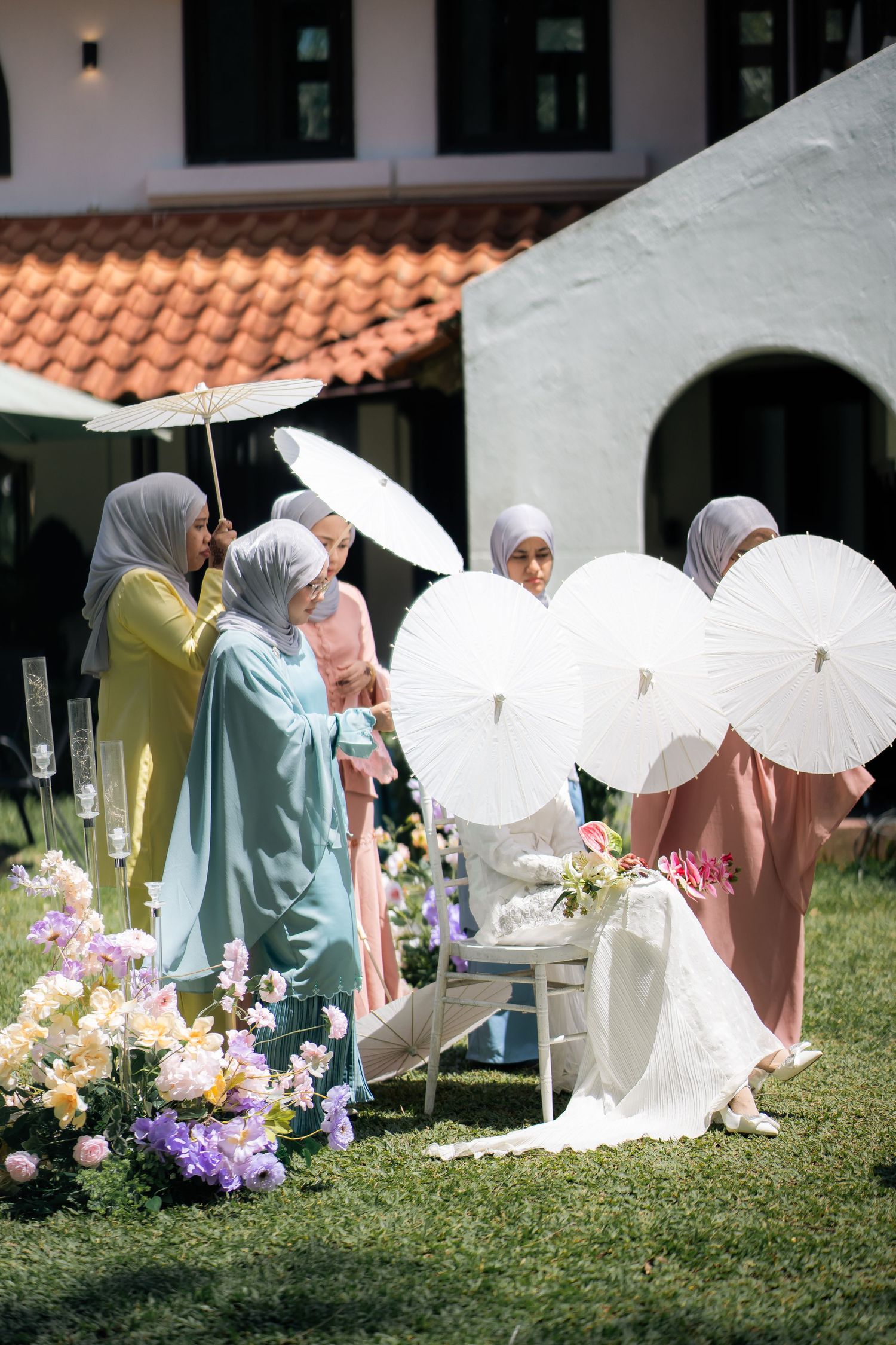 Wedding guests with white parasols gather on a grassy lawn with colorful floral arrangements and terracotta roofing.